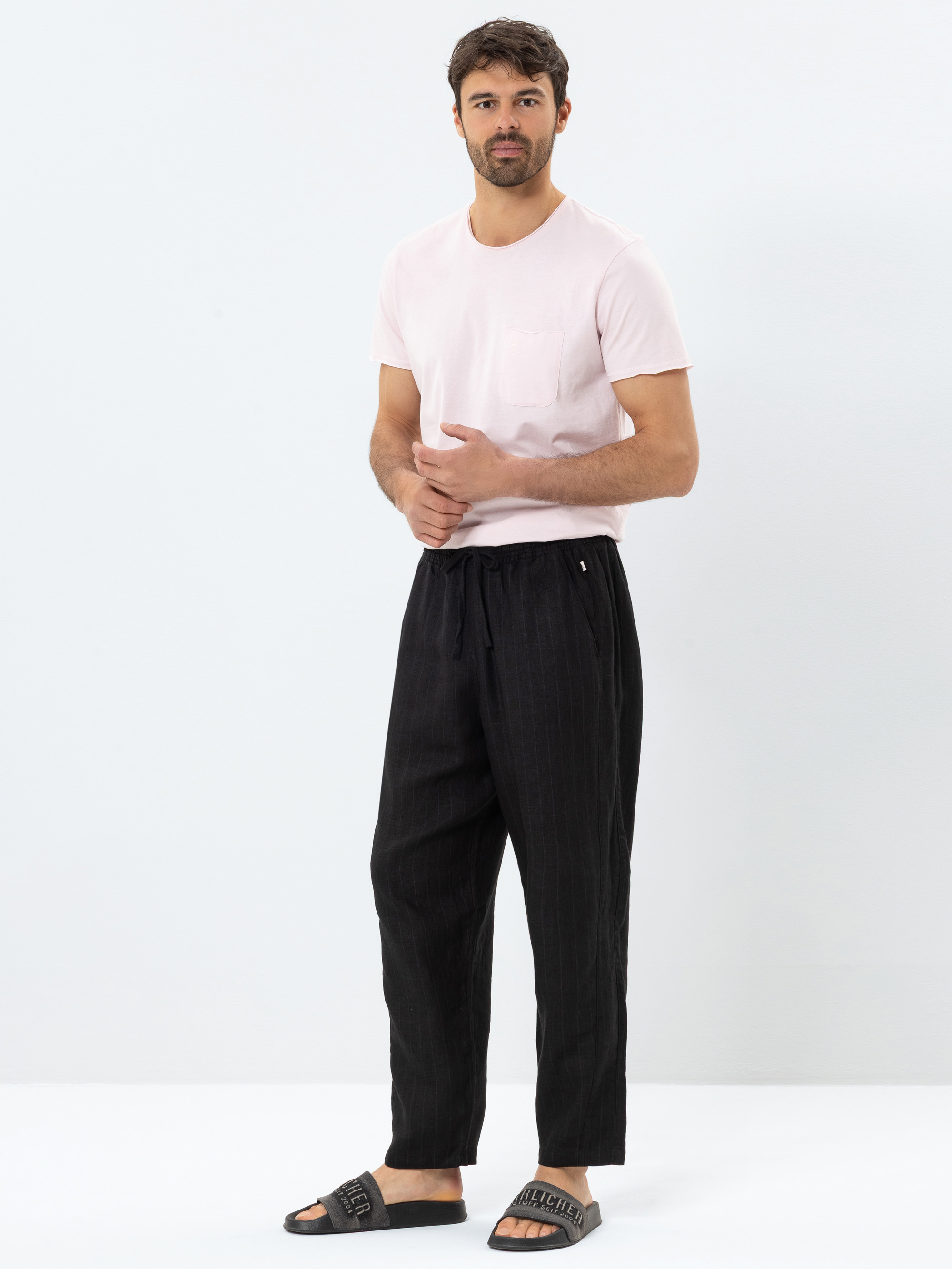 Ein Mann steht vor einem weißen Hintergrund und trägt ein hellrosa T-Shirt, eine weite schwarze Hose mit Kordelzug und schwarze Slide-Sandalen. Er blickt leicht zur Seite und hat die Hände sanft umklammert.