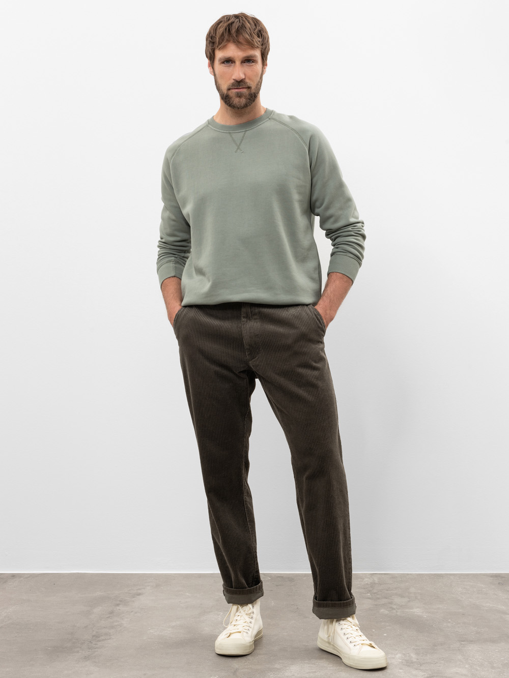 Ein Mann steht vor einem schlichten weißen Hintergrund, trägt ein salbeigrünes Sweatshirt, eine dunkelbraune Cordhose und weiße Turnschuhe, hat die Hände in den Taschen und schaut in die Kamera.