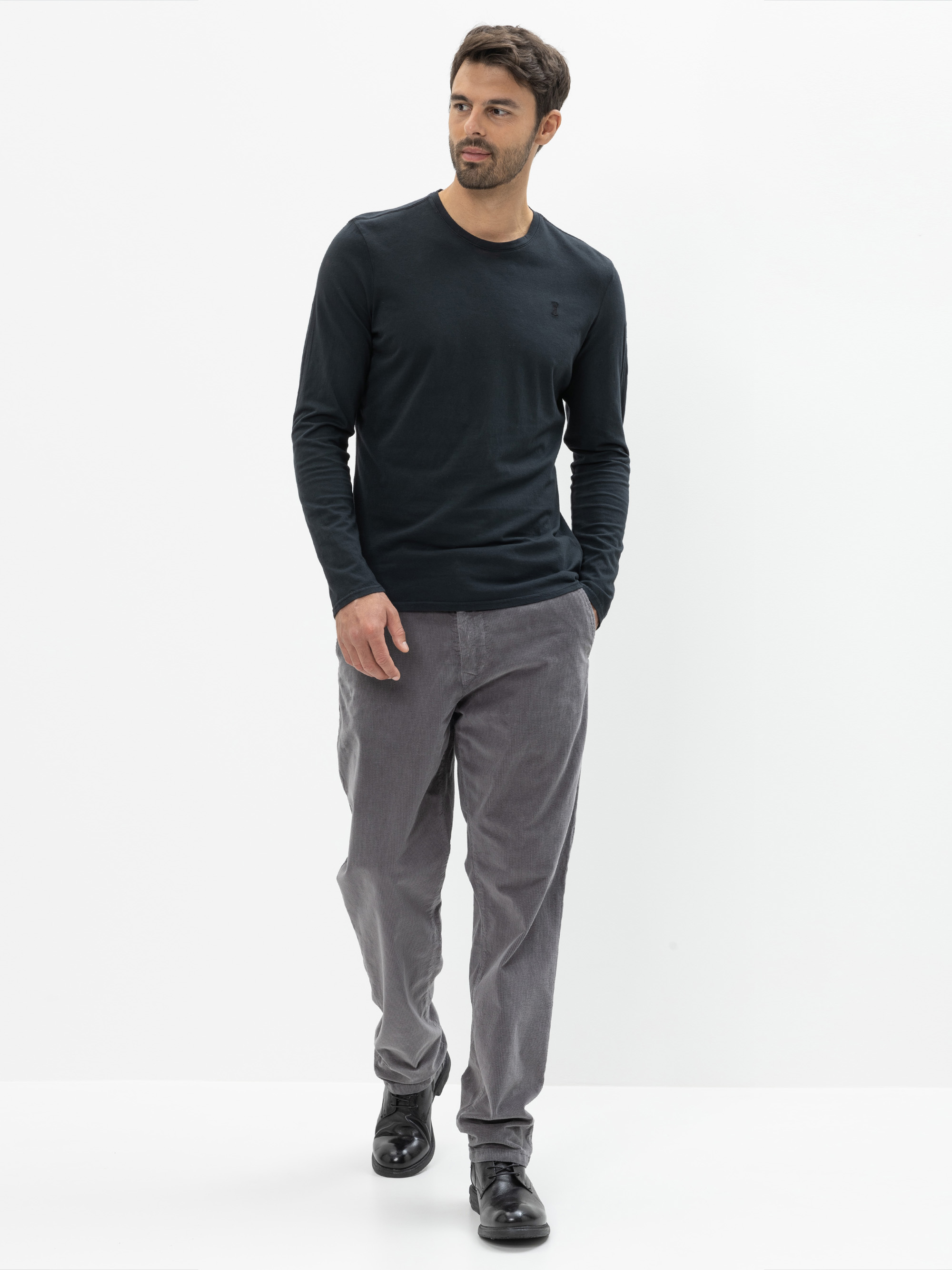 Ein Mann mit dunklem Haar und Bart geht in einem schwarzen, langärmeligen Hemd, einer hellgrauen Hose und schwarzen Schuhen vor einem schlichten weißen Hintergrund.