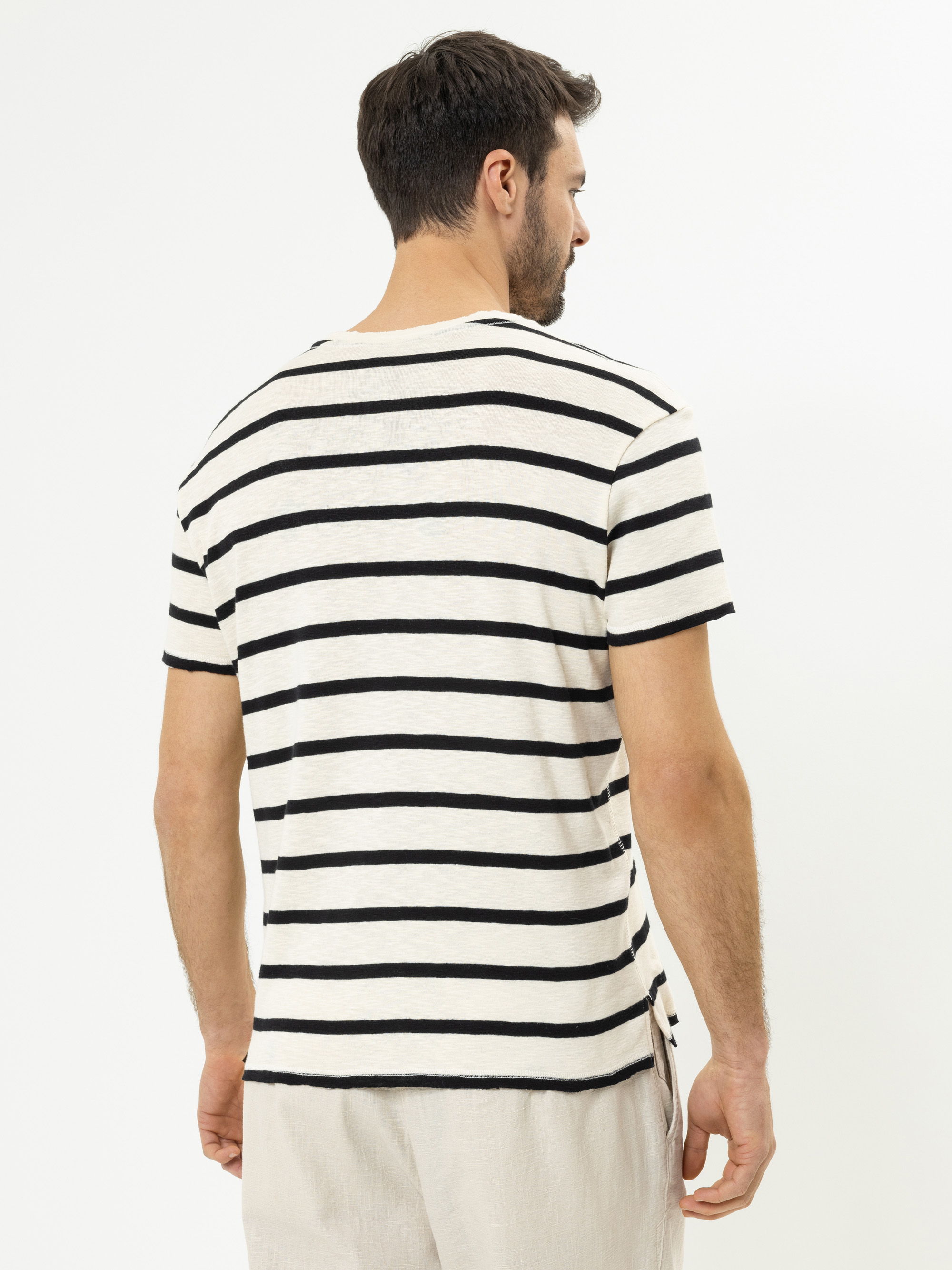Ein Mann steht mit dem Rücken zur Kamera, trägt ein weißes T-Shirt mit schwarzen Querstreifen und eine hellbeige Hose vor einem schlichten weißen Hintergrund.