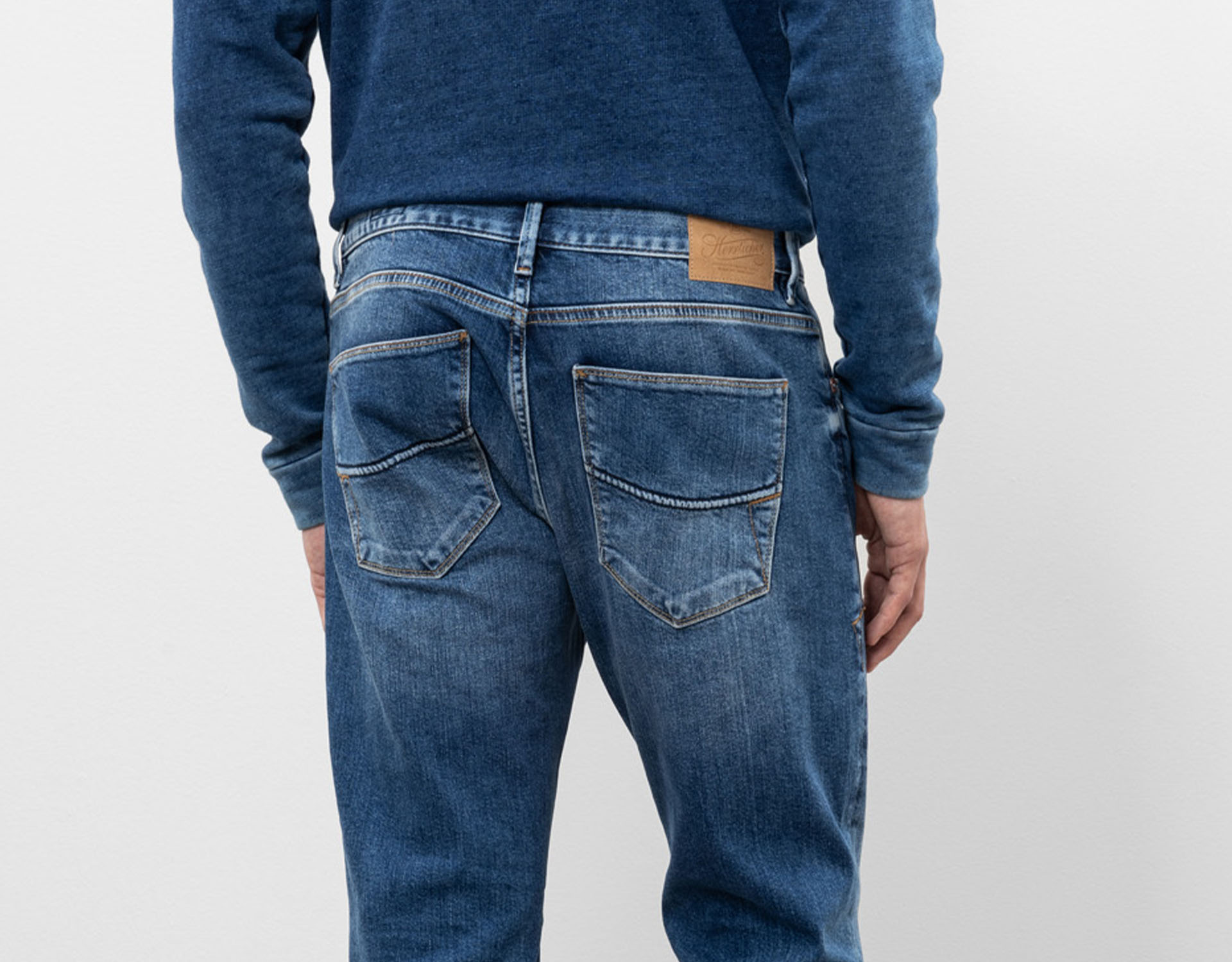 Das Bild zeigt die Rückenansicht einer Person, die ein blaues Sweatshirt und eine blaue Denim-Jeans mit sichtbaren Gesäßtaschen und einem braunen Lederaufnäher am Bund trägt. Der Hintergrund ist einfarbig und hell gefärbt.