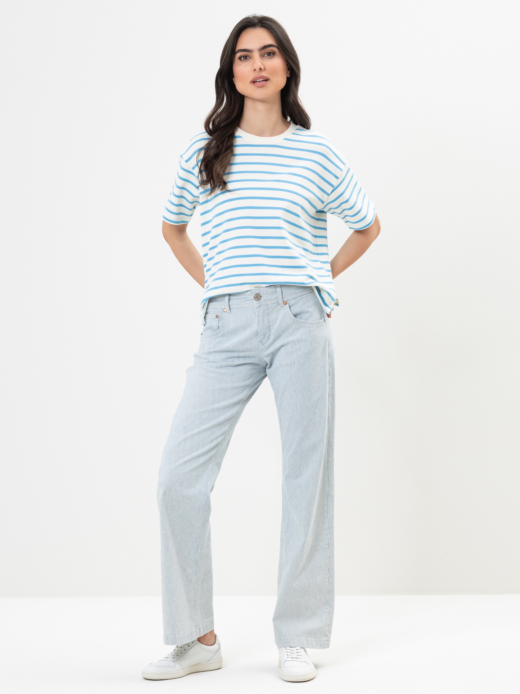 Eine Frau steht vor einem schlichten weißen Hintergrund und trägt ein blau-weiß gestreiftes kurzärmeliges Hemd, hellblaue Jeans mit weitem Bein und weiße Turnschuhe. Sie hat langes dunkles Haar und posiert mit einer Hand in der Tasche.