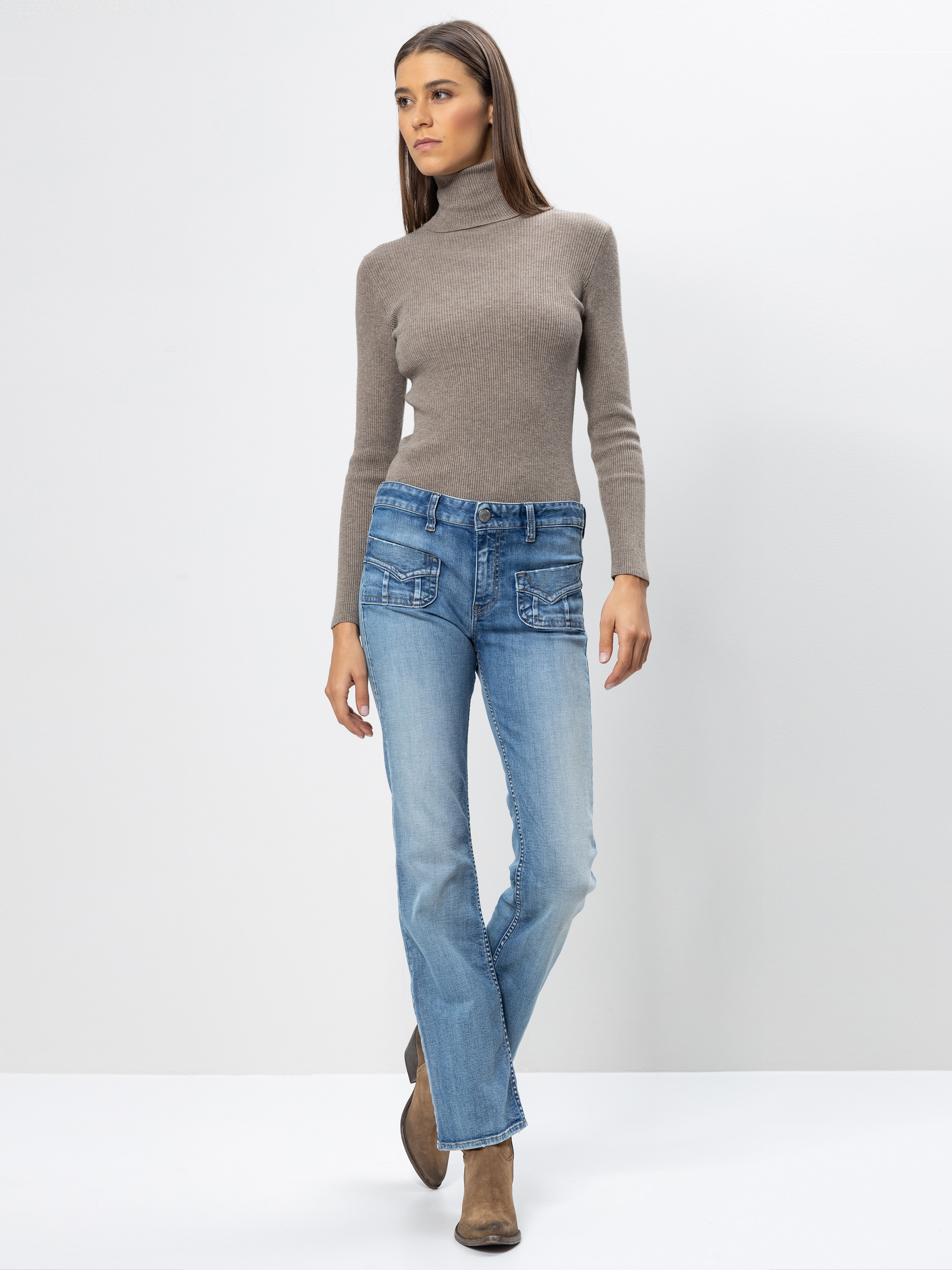 Eine Frau, die einen taupefarbenen Rollkragenpullover, hellblaue Jeans mit Vordertaschen und braune Stiefel trägt, steht vor einem schlichten weißen Hintergrund und schaut leicht zur Seite.