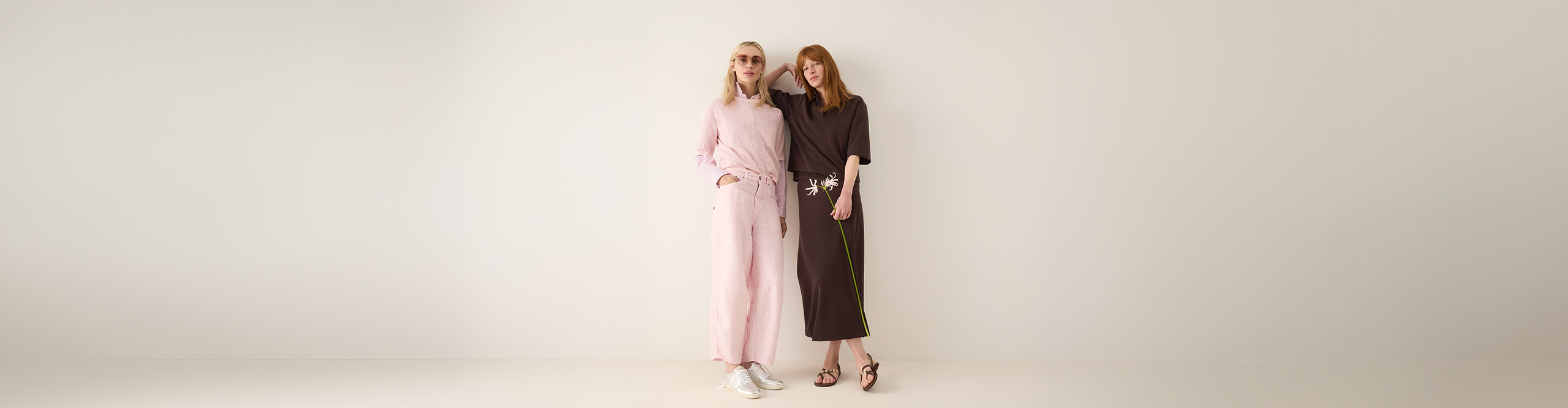 Zwei Frauen stehen zusammen vor einem schlichten, hellen Hintergrund. Die eine trägt einen hellrosa Overall mit weißen Turnschuhen, die andere ein dunkelbraunes Kleid mit Sandalen. Sie lächeln und posieren lässig.