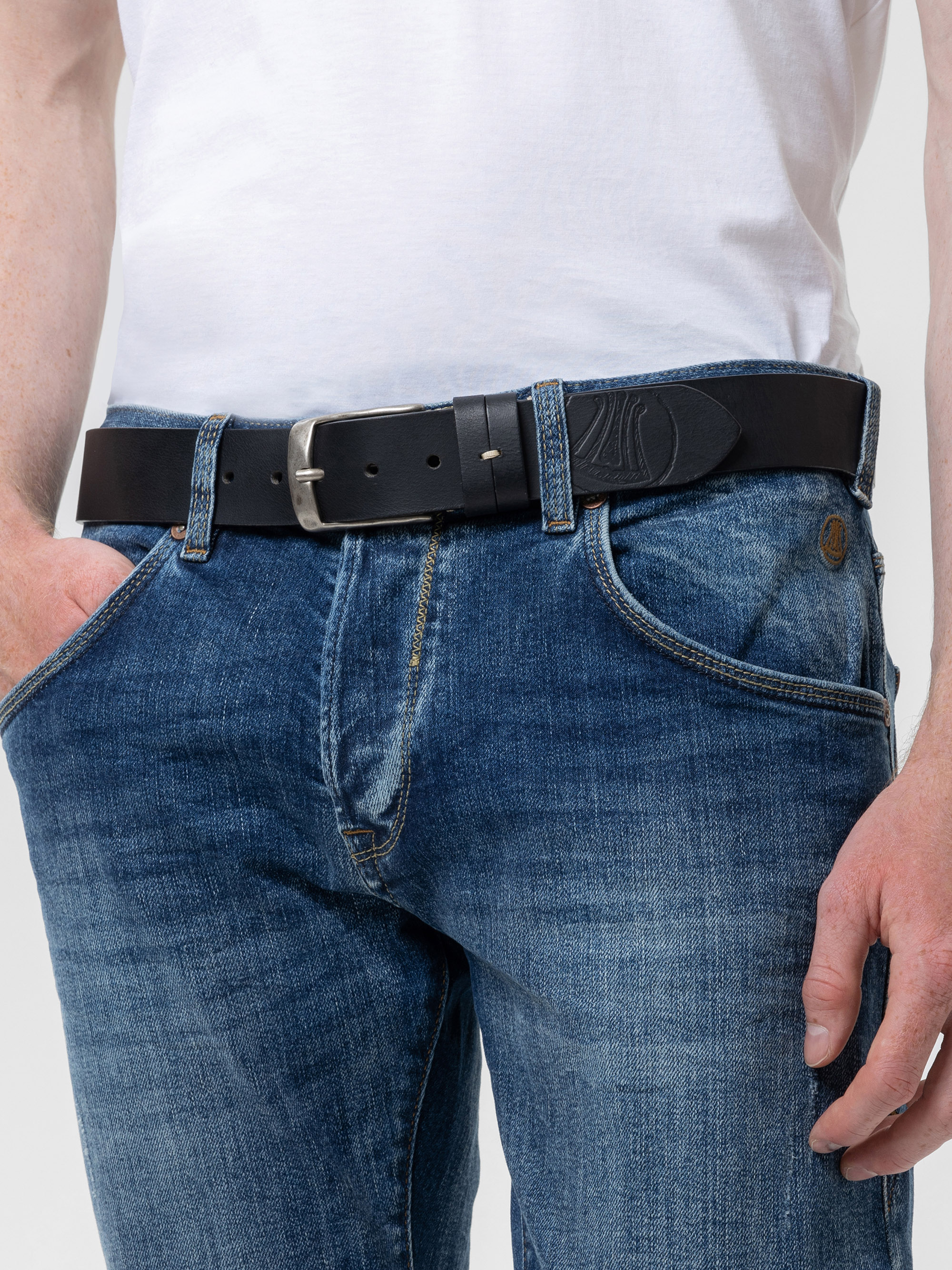 Eine Person trägt ein weißes T-Shirt, eine blaue Jeans und einen schwarzen Ledergürtel mit einer silbernen Schnalle. Eine Hand steckt in einer Tasche, die andere ruht an der Seite. Das Bild konzentriert sich auf den Taillenbereich.