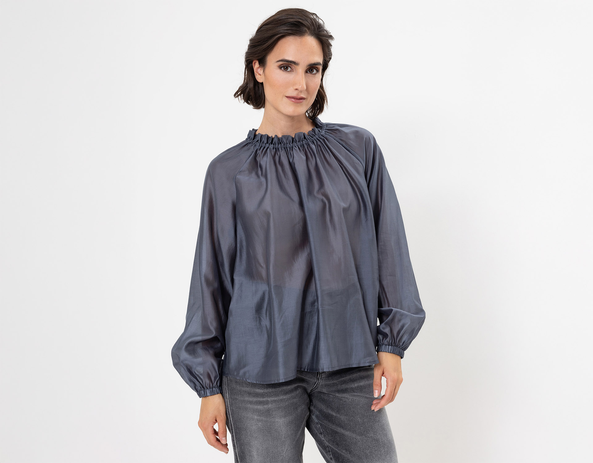 Eine Frau mit kurzen braunen Haaren trägt eine lockere, langärmelige, glänzende graue Bluse mit gerüschtem Ausschnitt und verblichene graue Jeans vor einem schlichten weißen Hintergrund.