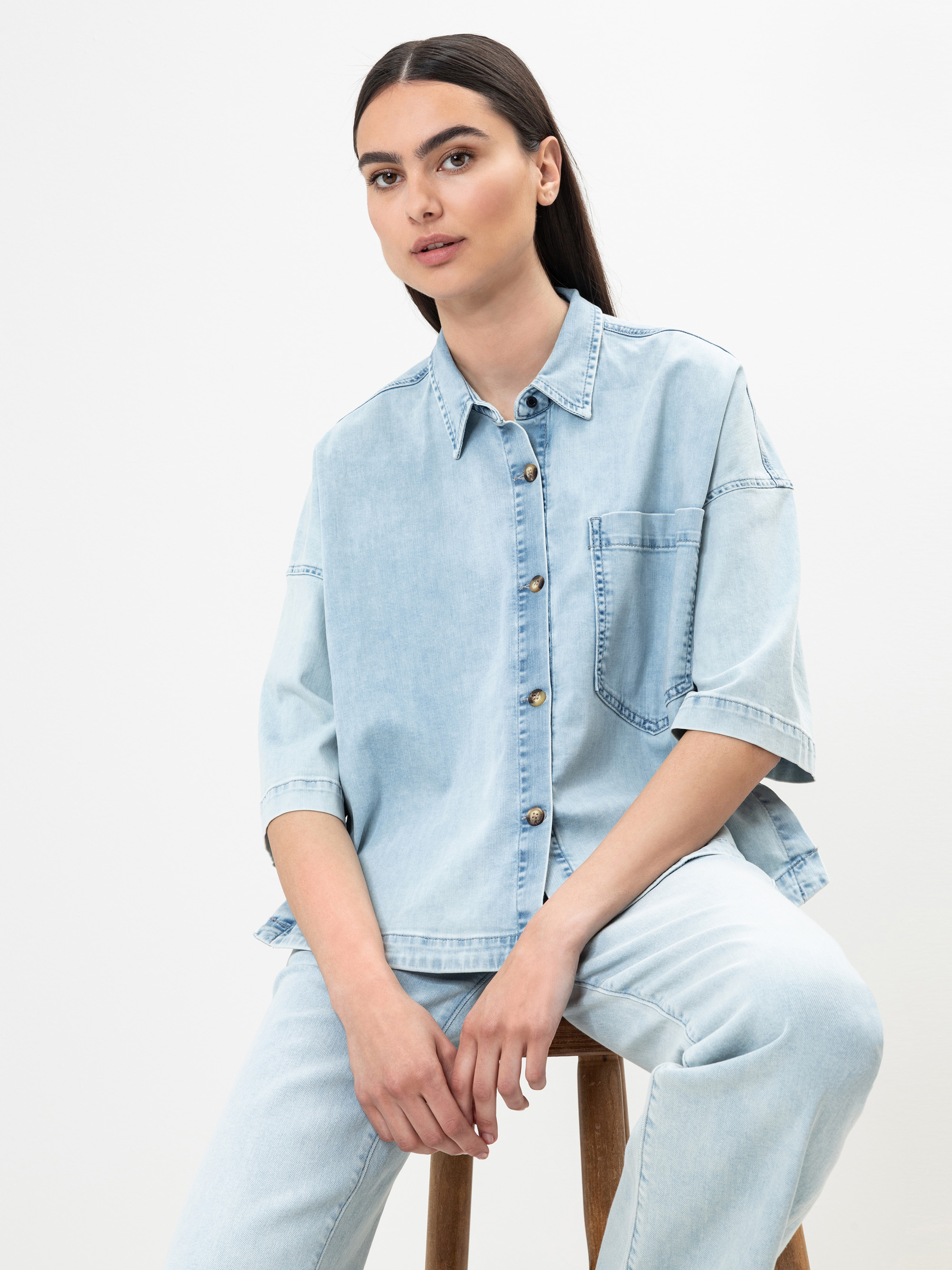 Eine Frau mit langen dunklen Haaren sitzt auf einem Hocker und trägt ein hellblaues Jeanshemd mit kurzen Ärmeln und dazu passende hellblaue Jeans. Sie blickt leicht nach links vor einem schlichten weißen Hintergrund.
