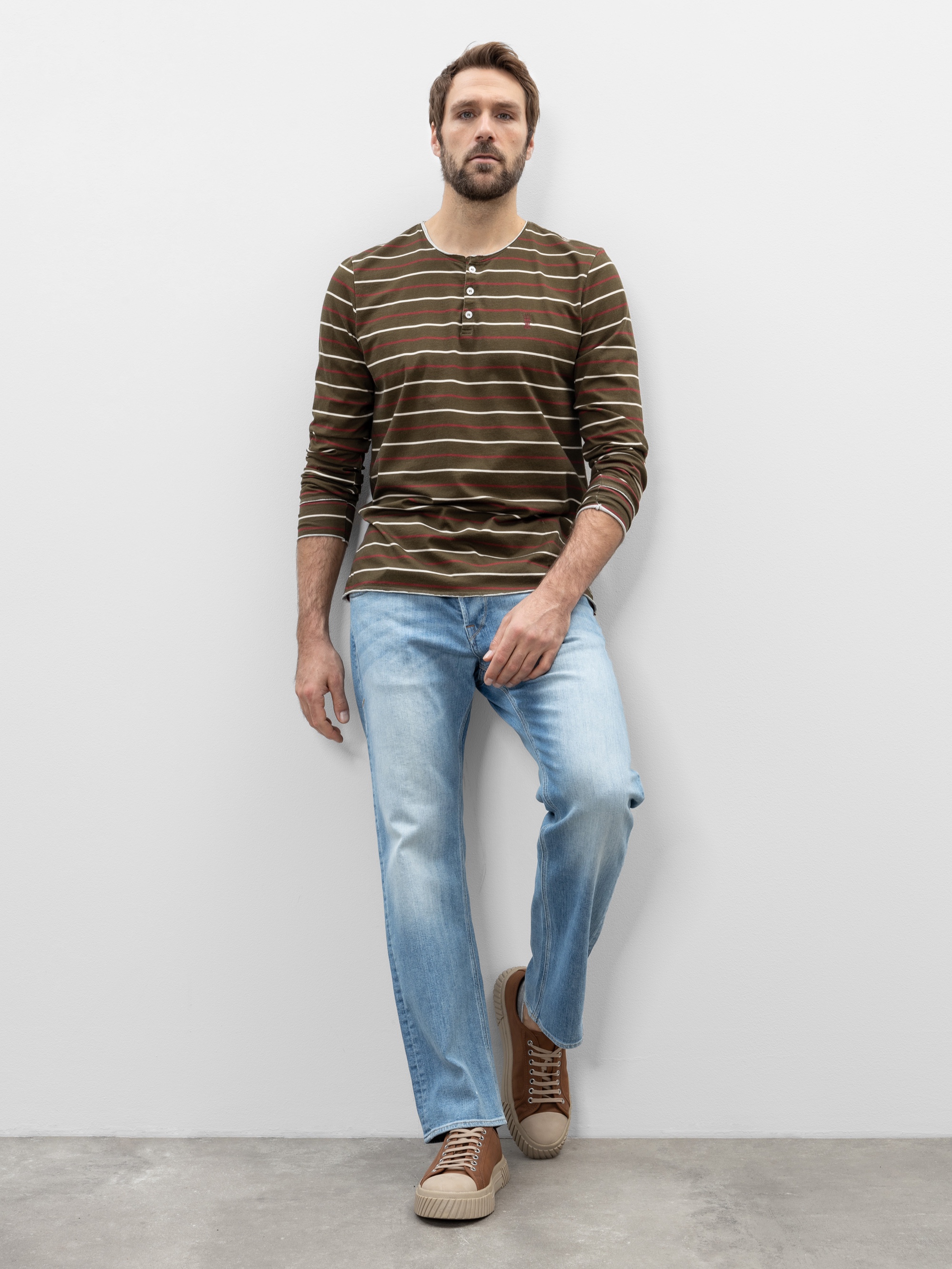 Ein Mann mit kurzen braunen Haaren und einem Bart steht vor einer weißen Wand. Er trägt ein braun-oliv gestreiftes langärmeliges Hemd, hellblaue Jeans und braune Turnschuhe mit weißen Sohlen.