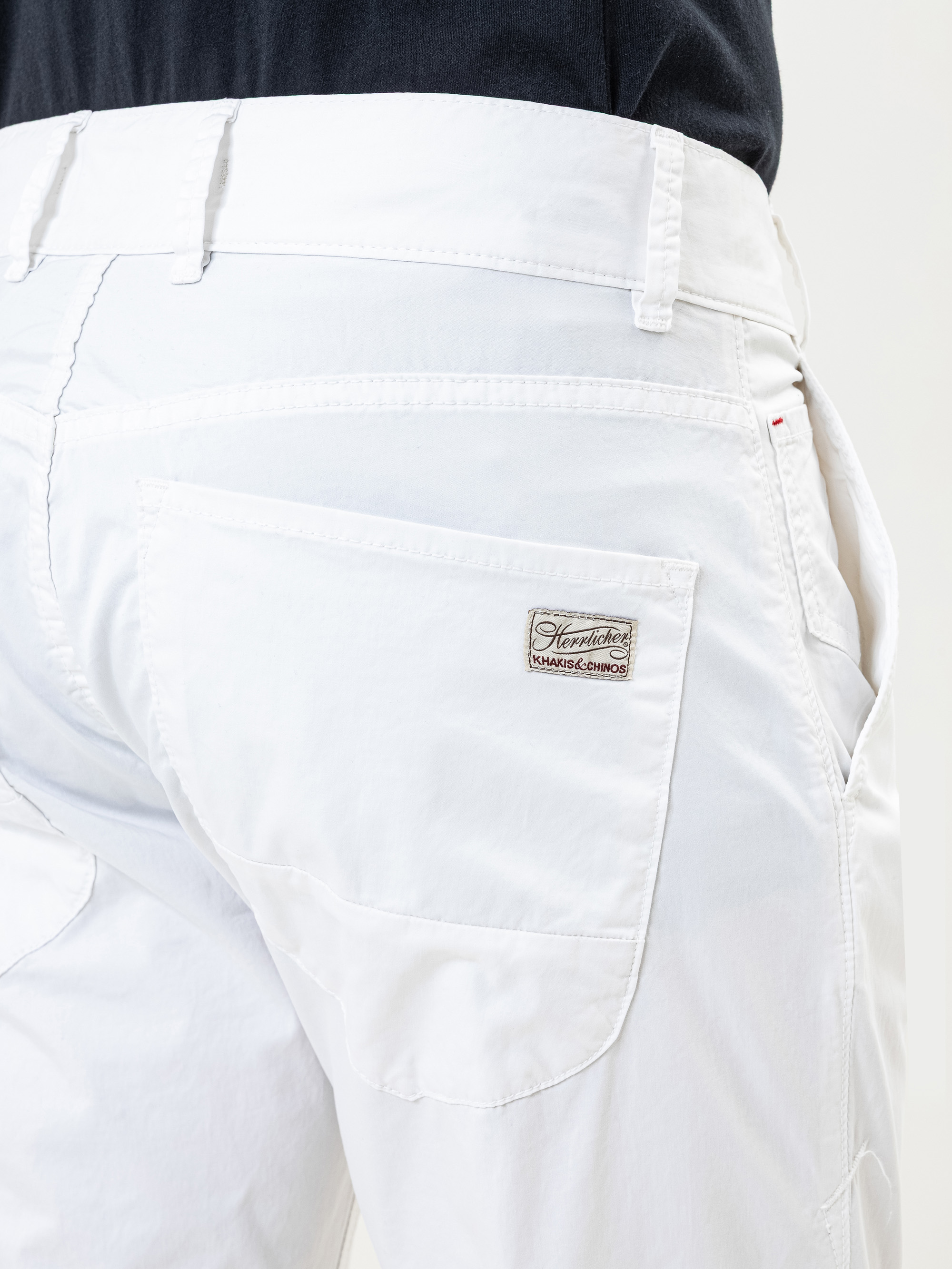 Nahaufnahme des Rückens einer Person, die eine weiße Hose mit einem sichtbaren Etikett mit der Aufschrift Stone Island auf der rechten Gesäßtasche trägt. Die Person trägt außerdem ein dunkles Hemd, das in die Hose gesteckt ist.