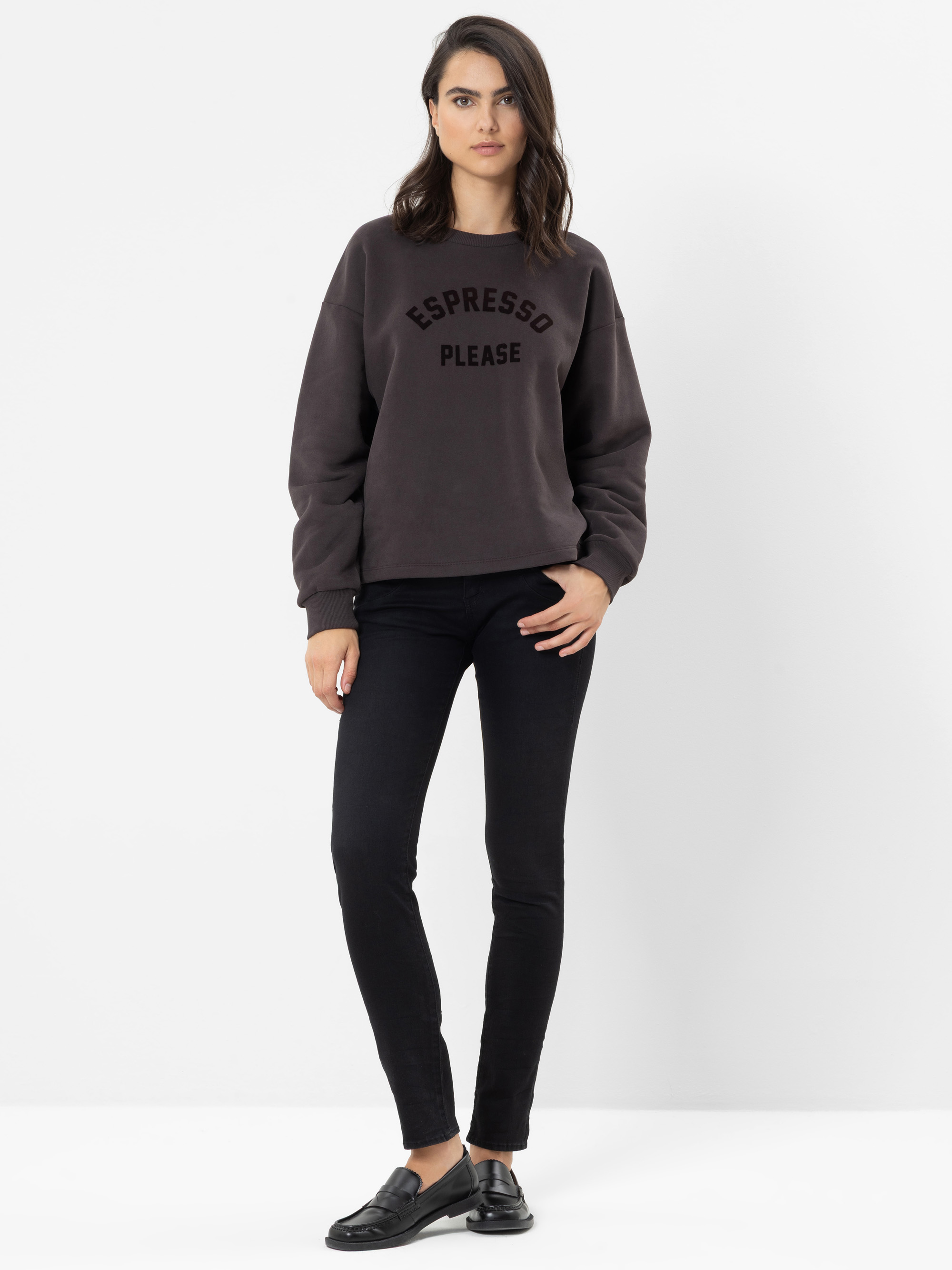 Eine Frau steht vor einem weißen Hintergrund und trägt ein dunkles Sweatshirt mit der Aufschrift „ESPRESSO PLEASE“, eine schwarze Skinny-Jeans und schwarze Loafer. Sie hat dunkles Haar und einen neutralen Gesichtsausdruck.