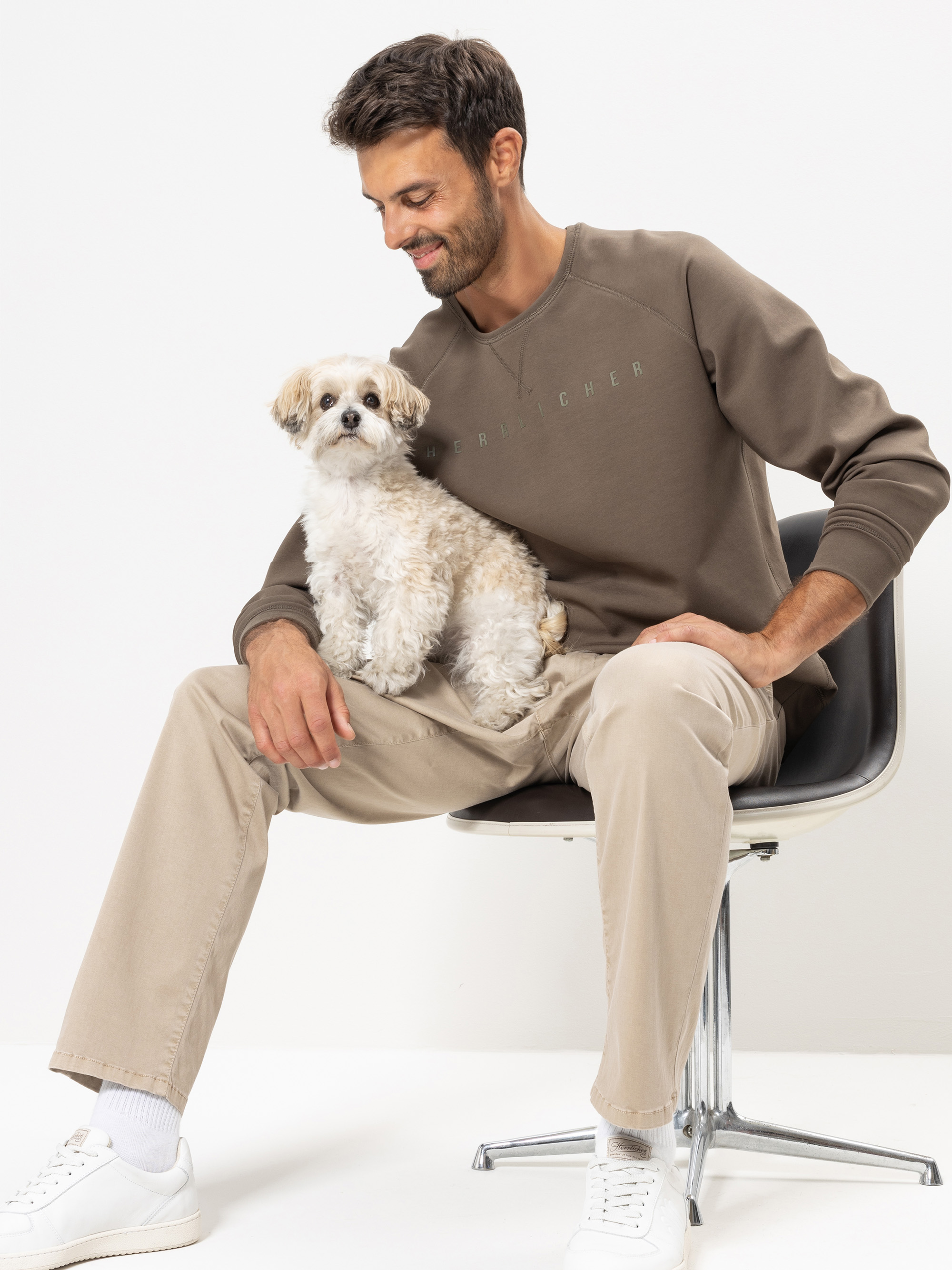 Ein Mann in einem braunen Sweatshirt und einer beigen Hose sitzt auf einem Stuhl und lächelt einem kleinen, flauschigen Hund zu, der auf seinem Schoß sitzt. Der Hintergrund ist einfarbig weiß.