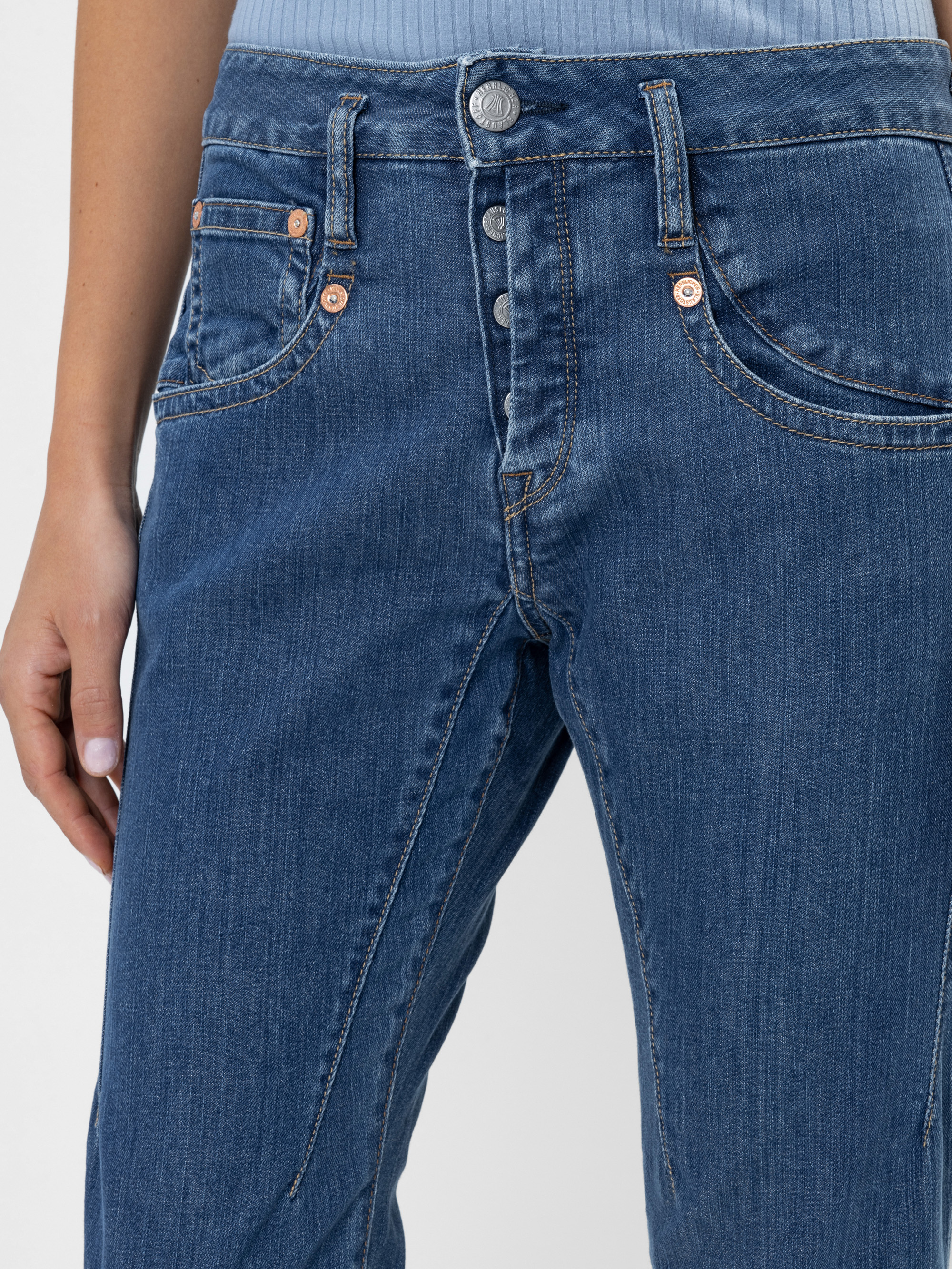 Eine Person, die eine blaue Jeans mit sichtbaren Knöpfen und Nähten trägt, steht mit einer Hand locker an der Seite. Das Oberteil ihres hellblauen Hemdes ist teilweise sichtbar.