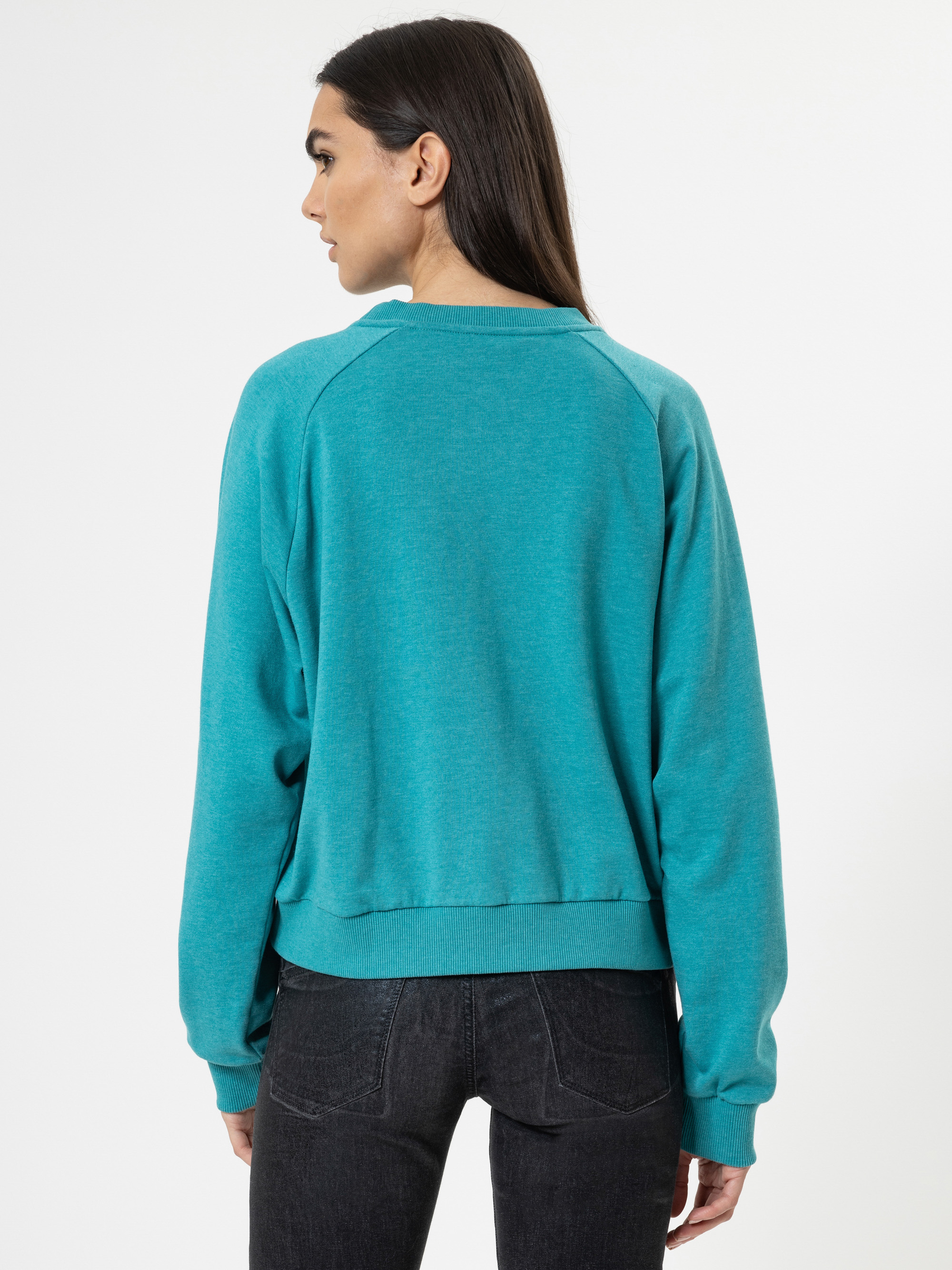 Eine Frau mit langen dunklen Haaren, die ein blaugrünes Sweatshirt und schwarze Jeans trägt, steht mit dem Gesicht zur Kamera vor einem schlichten weißen Hintergrund.