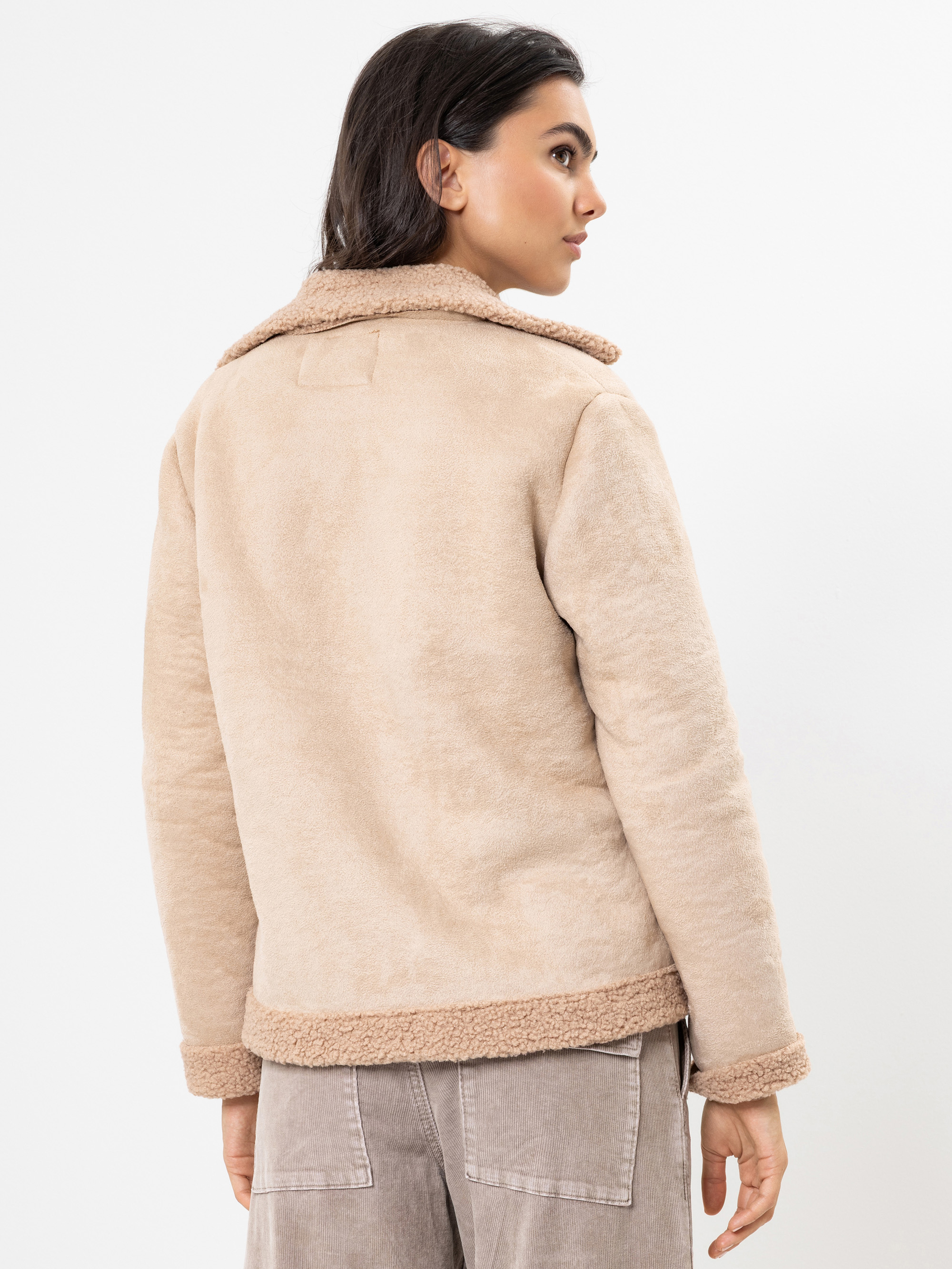Eine Frau mit dunklem Haar, die eine hellbeige, mit Shearling gefütterte Jacke und eine taupefarbene Hose trägt, steht mit dem Gesicht zur Kamera vor einem schlichten weißen Hintergrund.