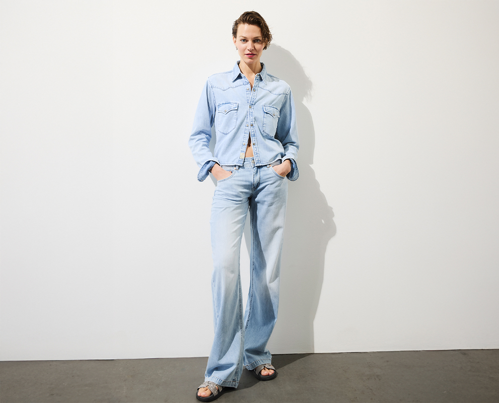 Eine Person steht an einer schlichten weißen Wand und trägt eine hellblaue Jeansjacke, dazu passende Jeans mit weitem Bein und Sandalen. Ihre Hände stecken in den Taschen und sie hat kurzes Haar.