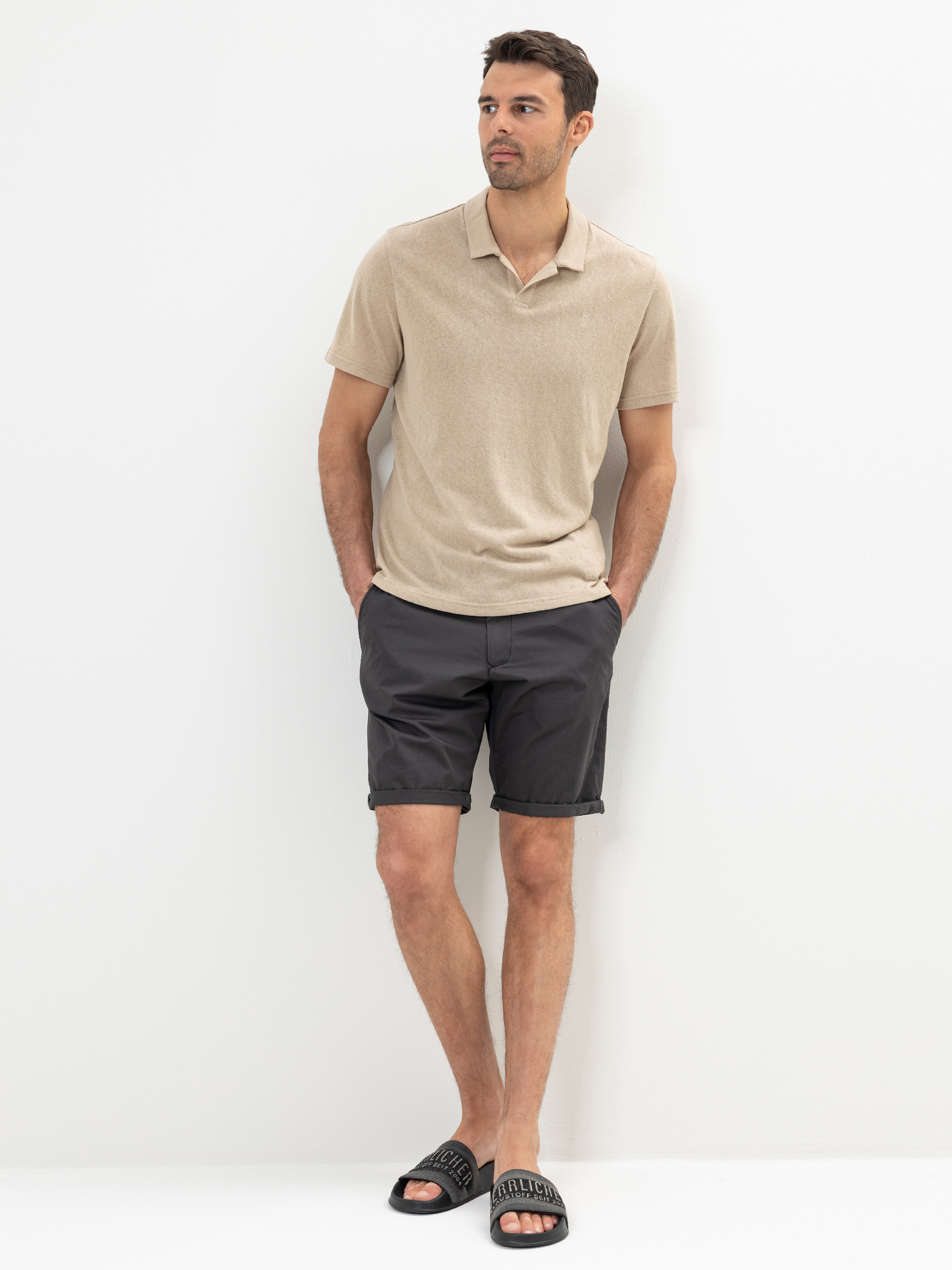 Ein Mann steht vor einer weißen Wand und trägt ein hellbeiges, kurzärmeliges Poloshirt, dunkelgraue Shorts und schwarze Slide-Sandalen. Er hat die Hände in den Hosentaschen und blickt mit neutraler Miene leicht zur Seite.