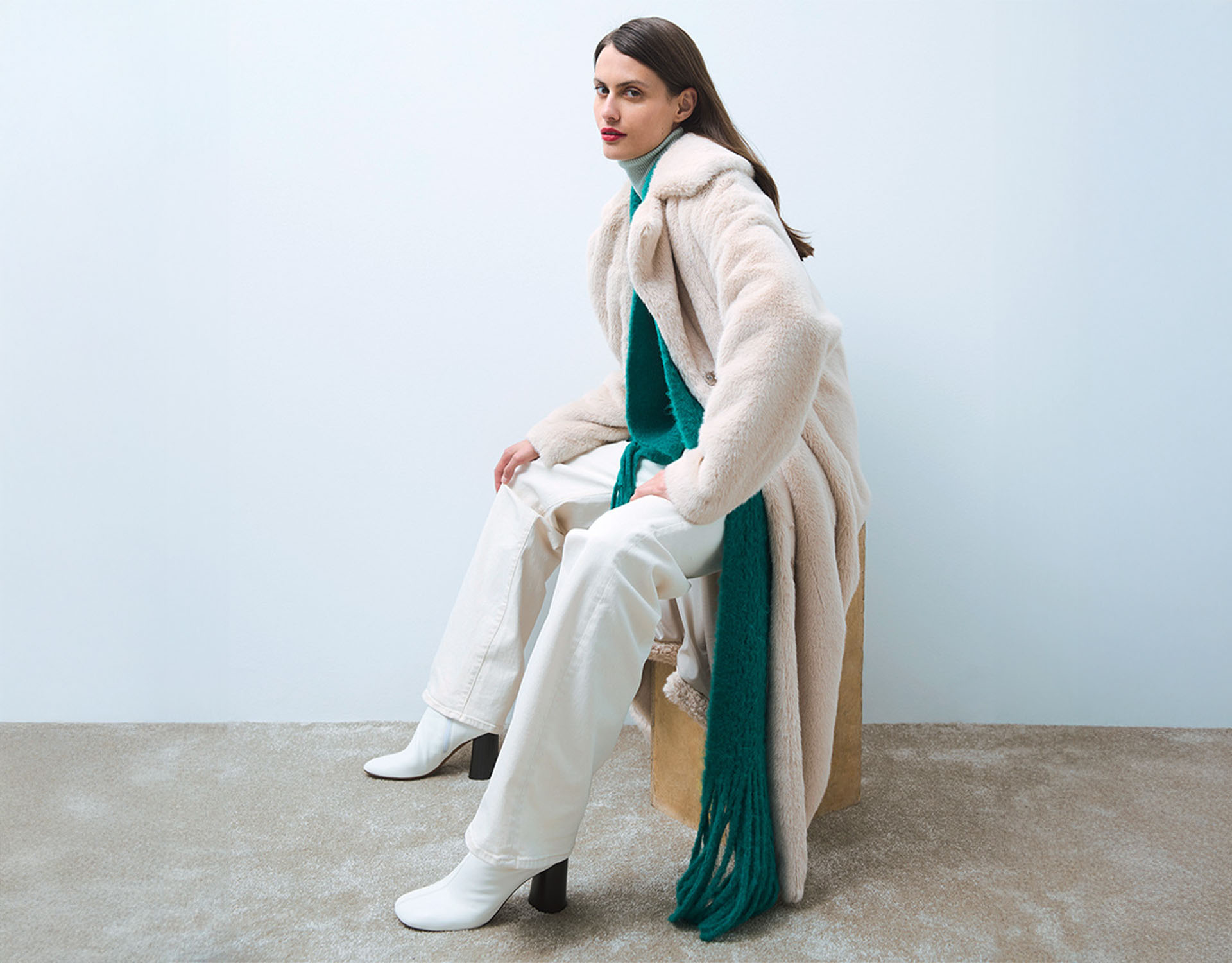 Eine Frau mit glattem braunem Haar sitzt auf einem Hocker und trägt einen langen cremefarbenen Mantel, eine weiße Hose, weiße Stiefel und einen blaugrünen Pullover mit einem passenden Schal vor einem schlichten hellen Hintergrund.