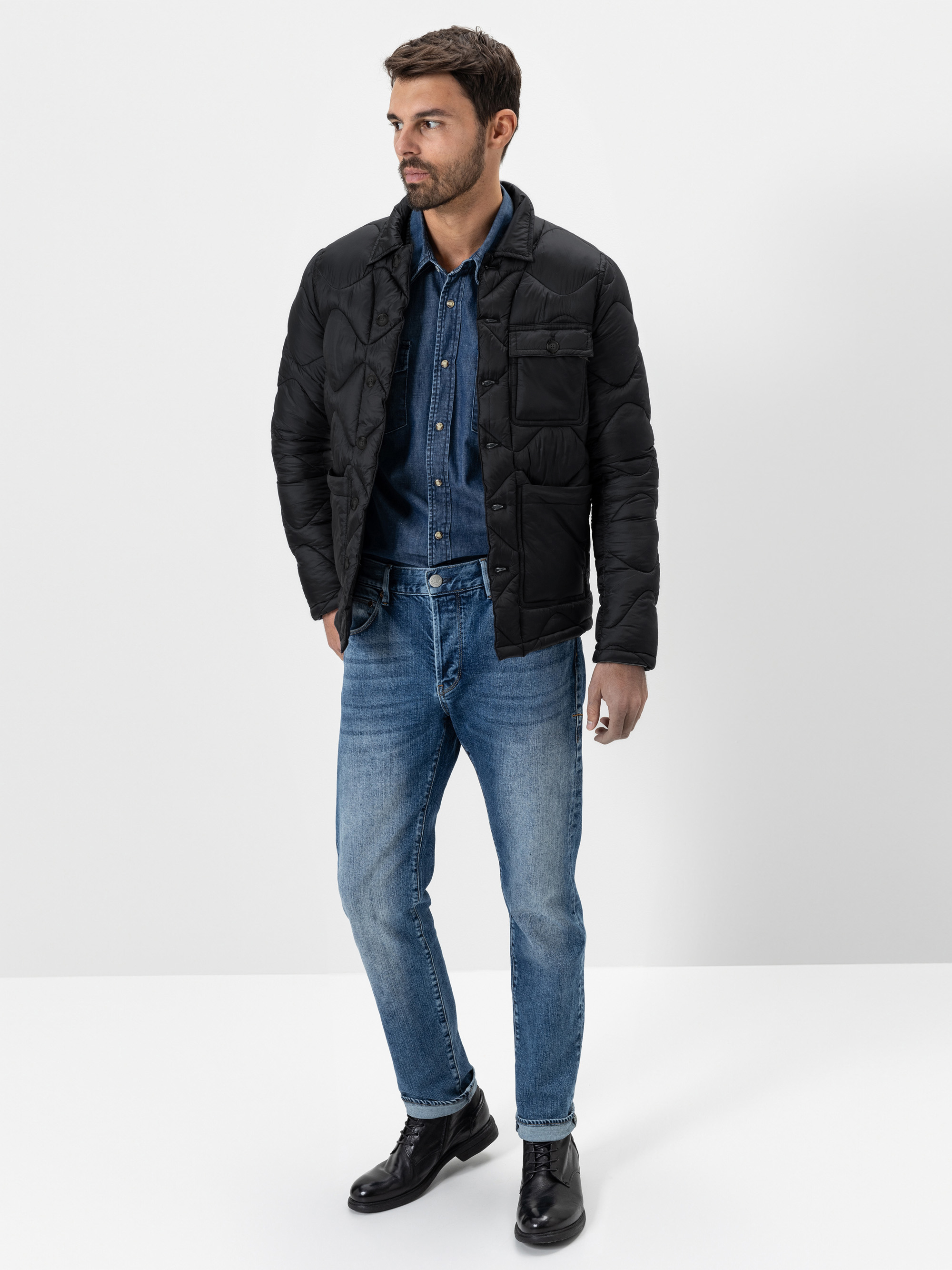 Ein Mann, der eine schwarze Pufferjacke über einem Jeanshemd und eine blaue Jeans, gepaart mit schwarzen Schuhen, trägt, steht vor einem schlichten weißen Hintergrund.