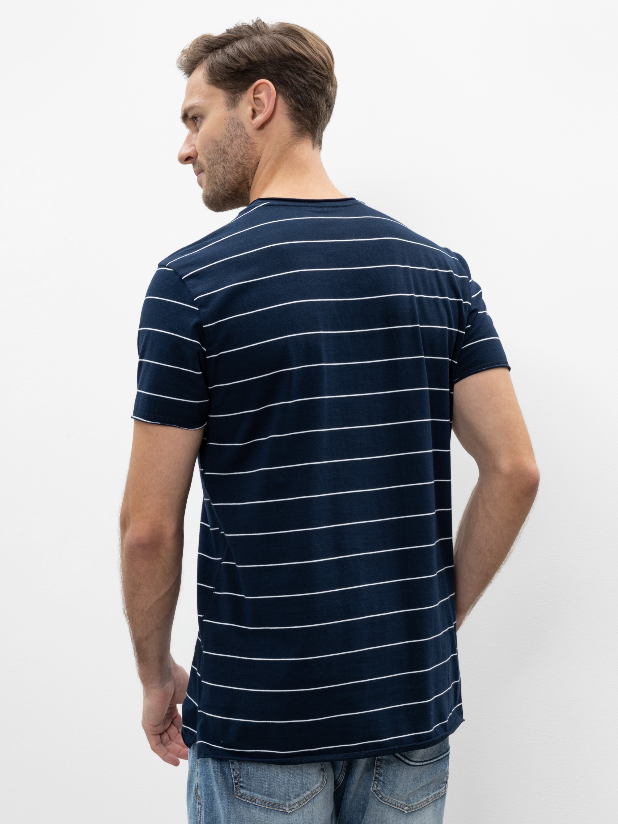 Ein Mann mit kurzen braunen Haaren, der ein marineblaues T-Shirt mit dünnen weißen Querstreifen und eine blaue Jeans trägt, steht mit dem Gesicht zur Kamera vor einem schlichten weißen Hintergrund.