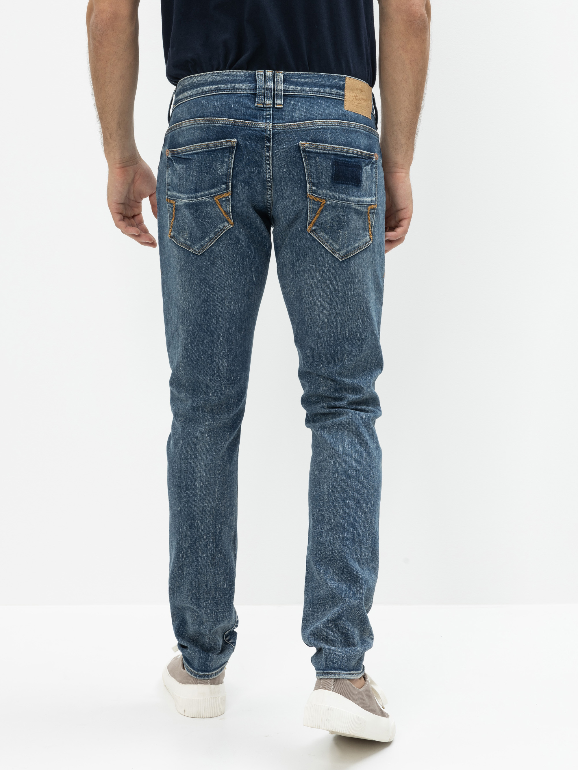 Eine Person, die eine blaue Jeans und weiße Turnschuhe trägt, steht mit dem Gesicht zur Seite und zeigt die Rückseite der Jeans vor einem schlichten weißen Hintergrund.