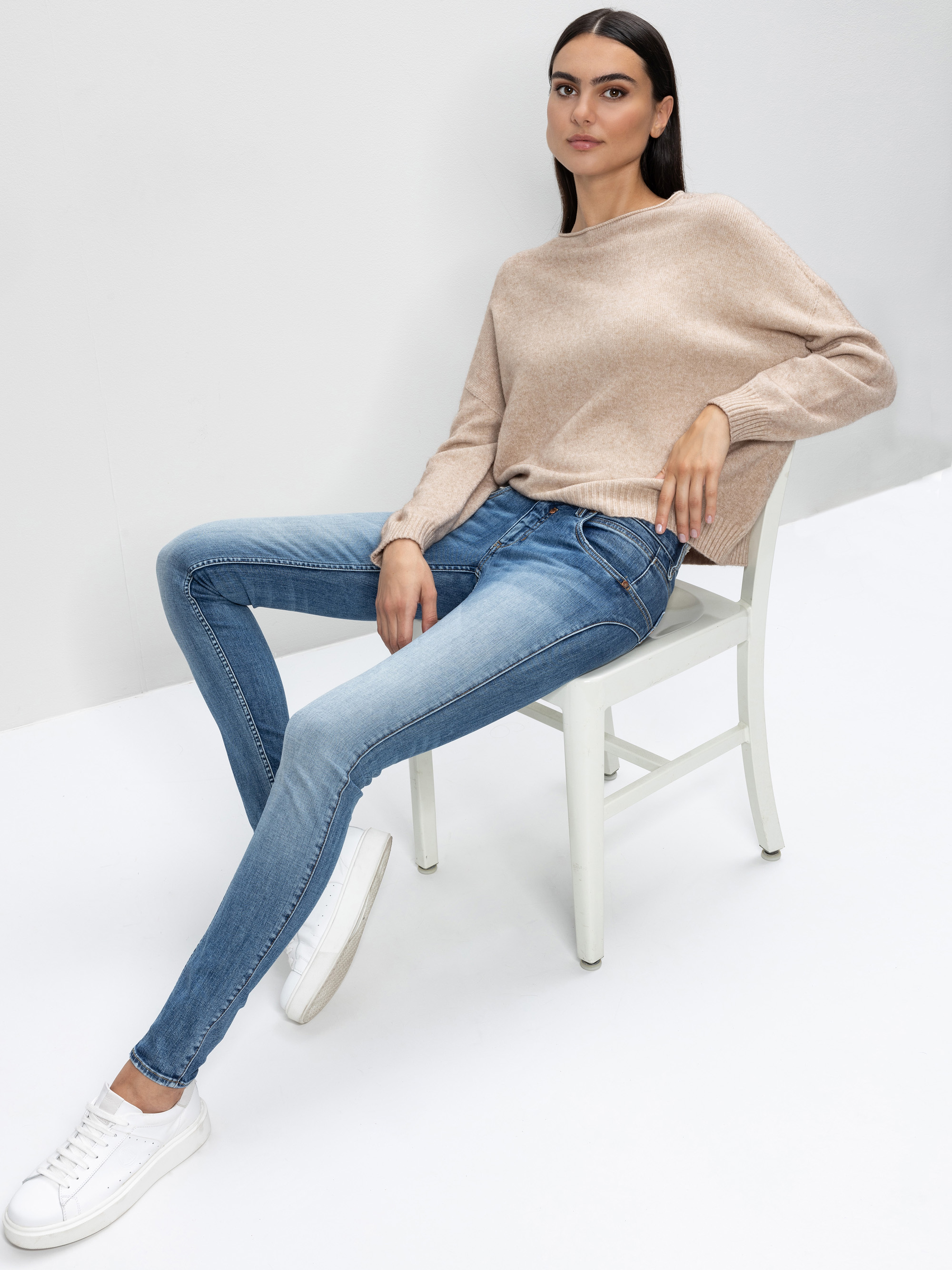 Eine Frau sitzt lässig auf einem weißen Stuhl, trägt einen beigen Pullover, blaue Röhrenjeans und weiße Turnschuhe, in einer minimalistischen, hellen Umgebung mit weißem Hintergrund.