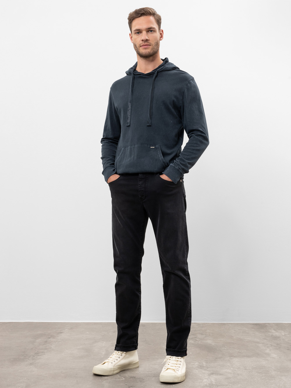 Ein Mann steht lässig vor einem schlichten weißen Hintergrund, trägt einen dunkelblauen Kapuzenpullover, schwarze Jeans und weiße Turnschuhe, die Hände in den Taschen und einen neutralen Gesichtsausdruck.