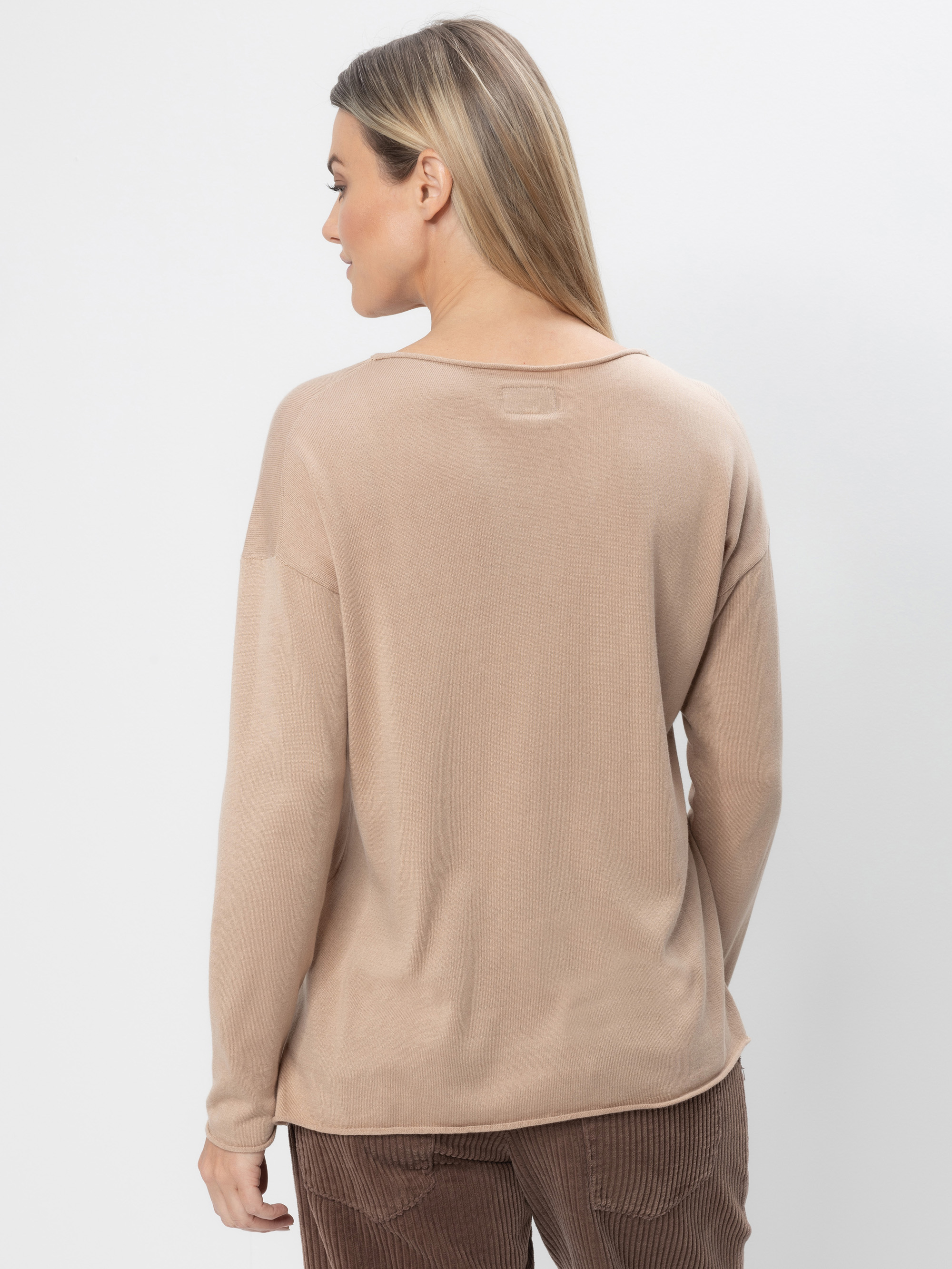 Eine Frau mit glattem, blondem Haar wird von hinten gezeigt. Sie trägt einen langärmeligen, hellbraunen Pullover und eine braune Cordhose und steht vor einem schlichten weißen Hintergrund.