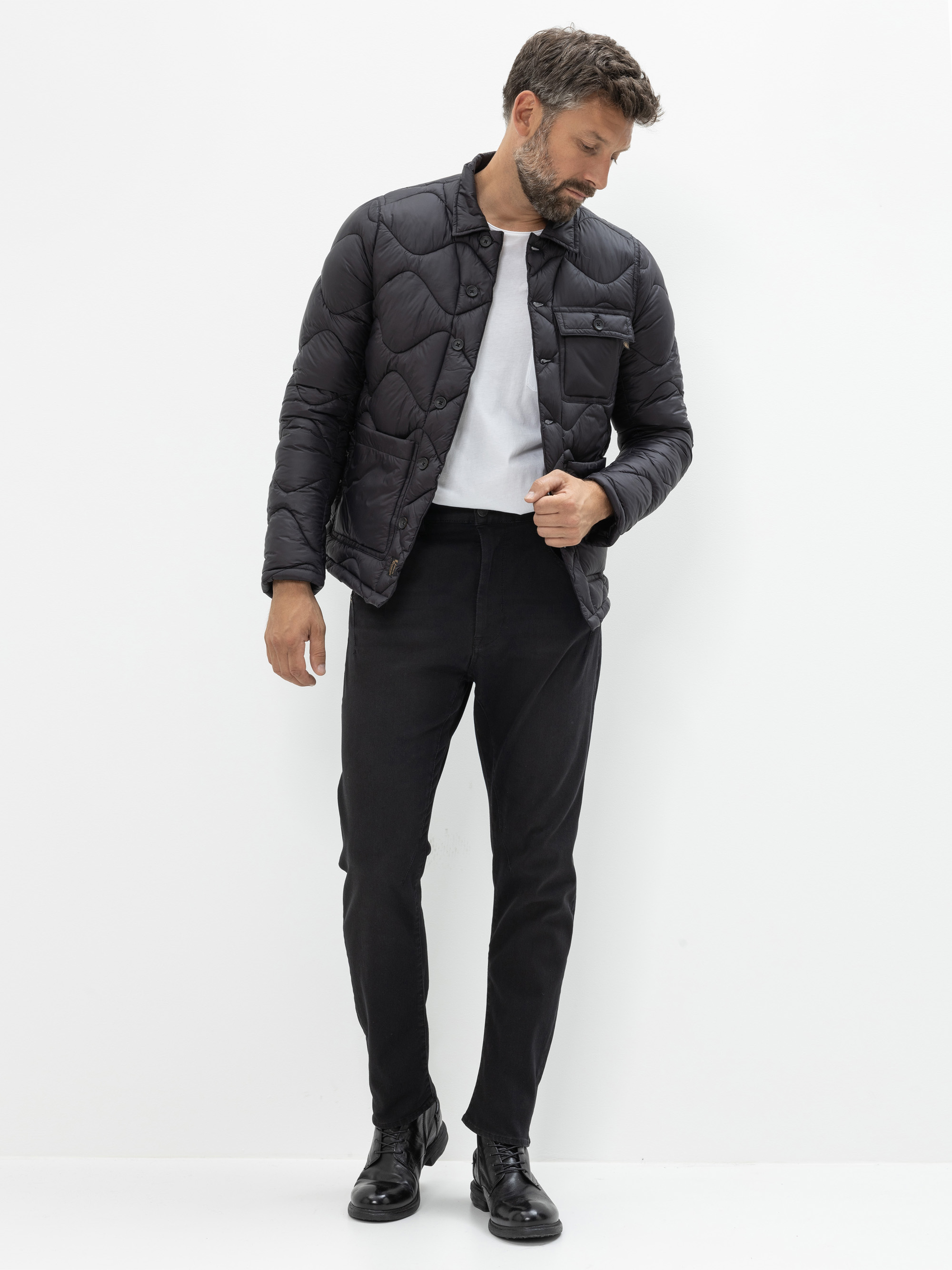 Ein Mann mit Bart trägt eine schwarze Steppjacke über einem weißen Hemd, eine dunkle Hose und schwarze Anzugschuhe. Er steht vor einem schlichten weißen Hintergrund und blickt nach links.