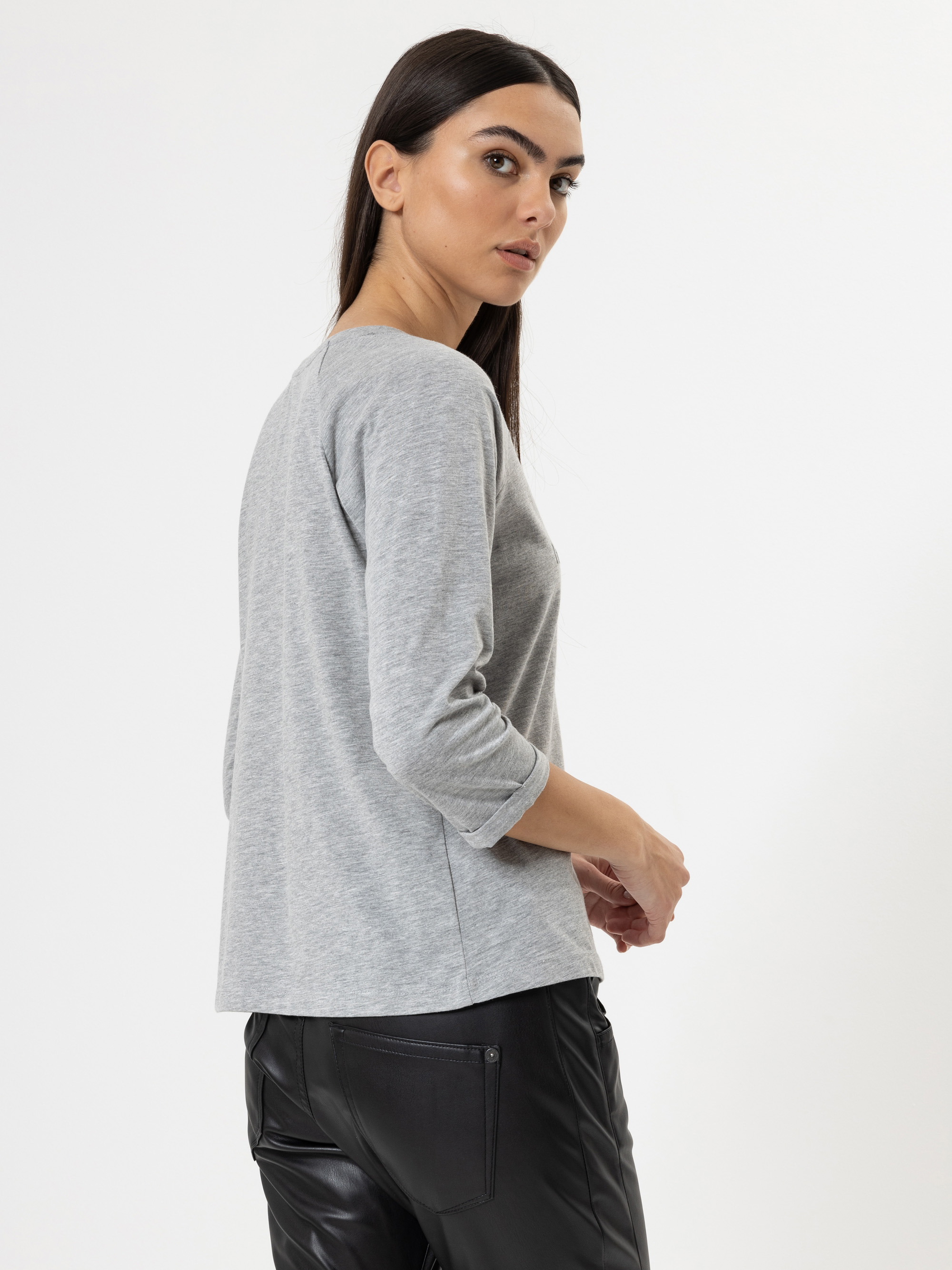 Eine Frau mit langen dunklen Haaren, bekleidet mit einem hellgrauen langärmeligen Oberteil und einer schwarzen Lederhose, steht vor einem schlichten weißen Hintergrund, leicht zur Seite gedreht und schaut über ihre Schulter.