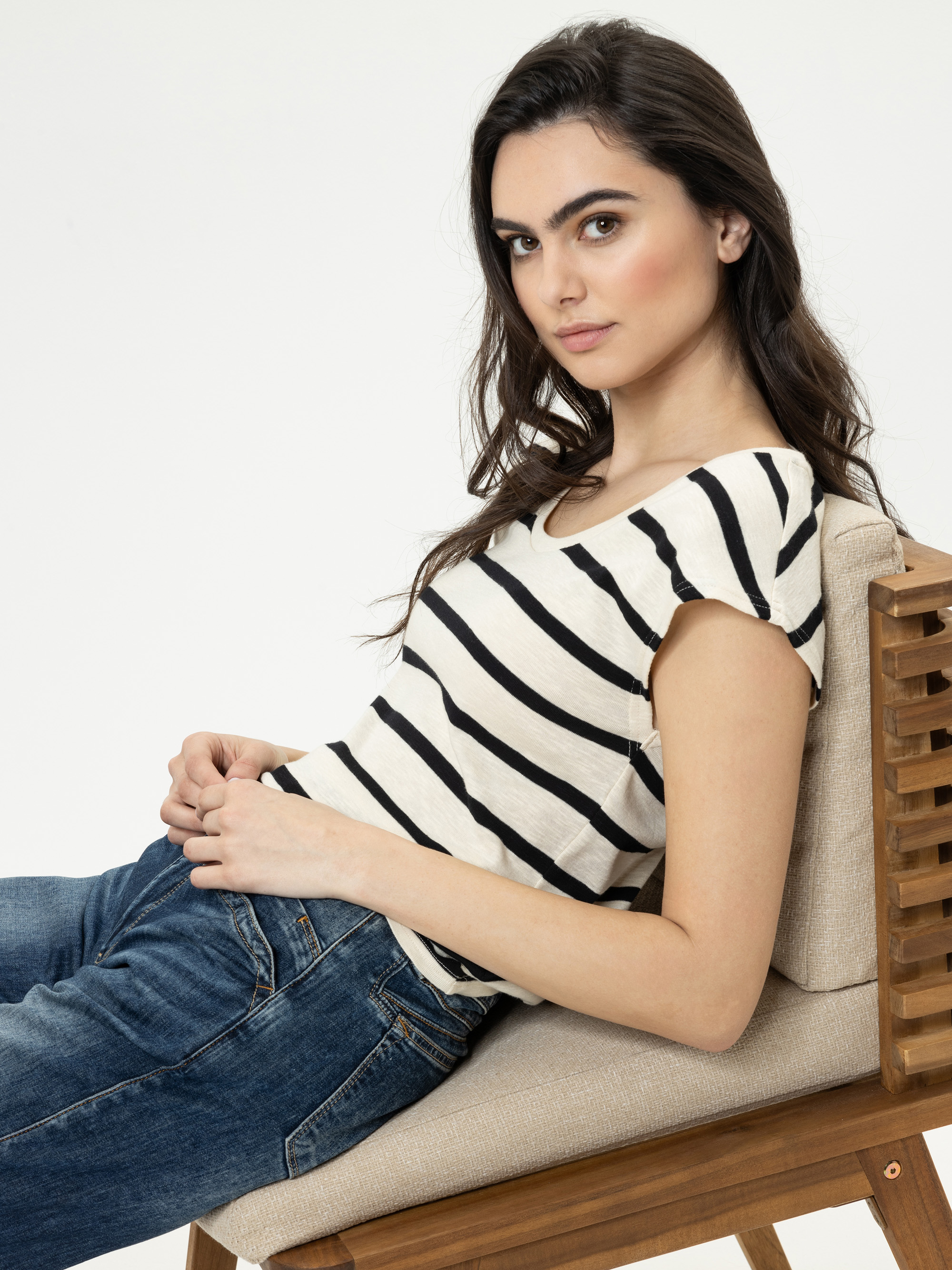 Eine Frau mit langen braunen Haaren, die ein schwarz-weiß gestreiftes Oberteil und eine blaue Jeans trägt, lehnt sich seitlich auf einem Holzstuhl zurück und blickt mit neutralem Blick in die Kamera vor einem schlichten weißen Hintergrund.