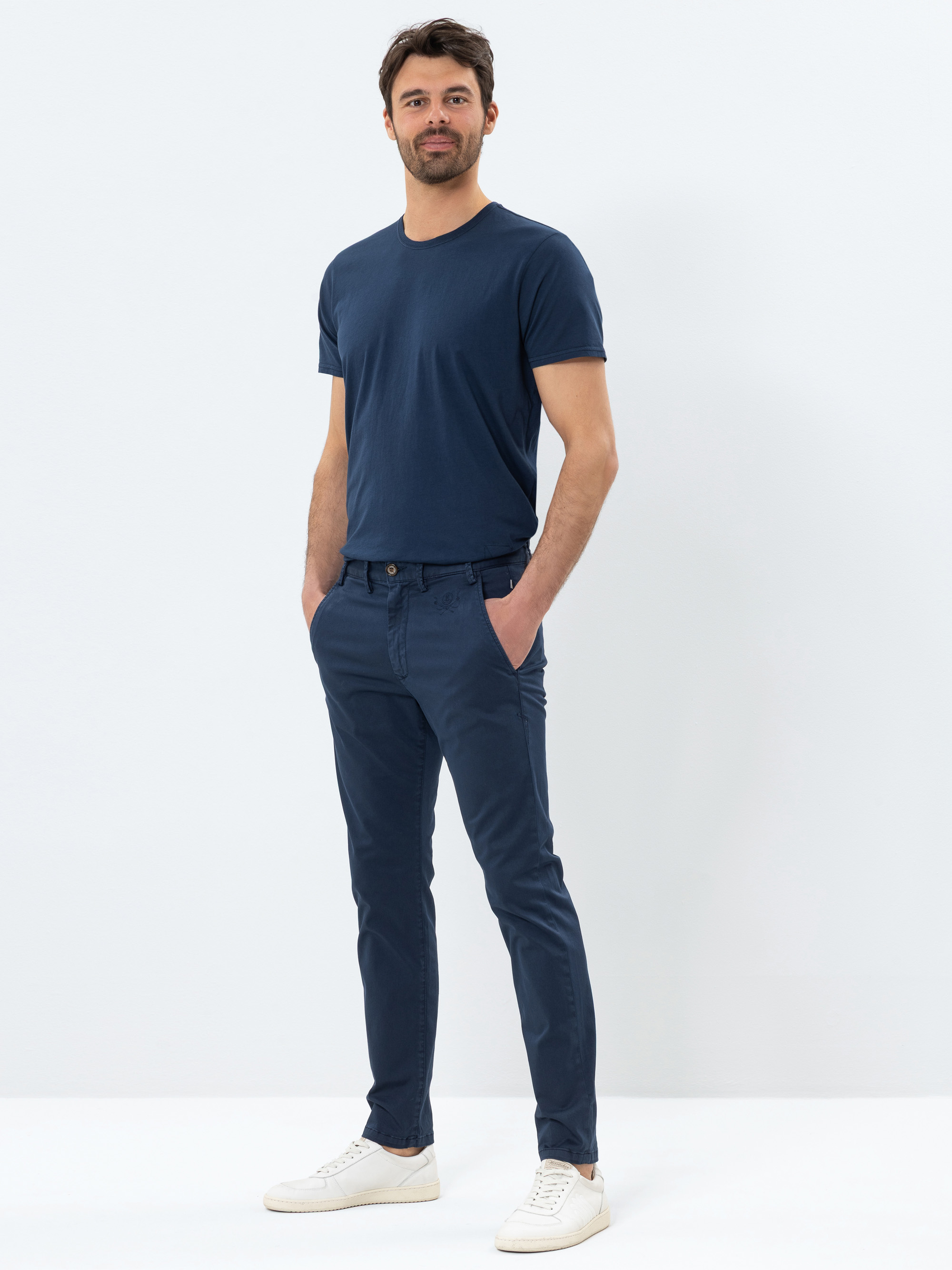 Ein Mann steht vor einem schlichten weißen Hintergrund und trägt ein marineblaues T-Shirt, eine blaue Slim-Fit-Hose und weiße Turnschuhe. Er nimmt eine entspannte Haltung ein, hat die Hände in den Taschen und schaut leicht lächelnd in die Kamera.