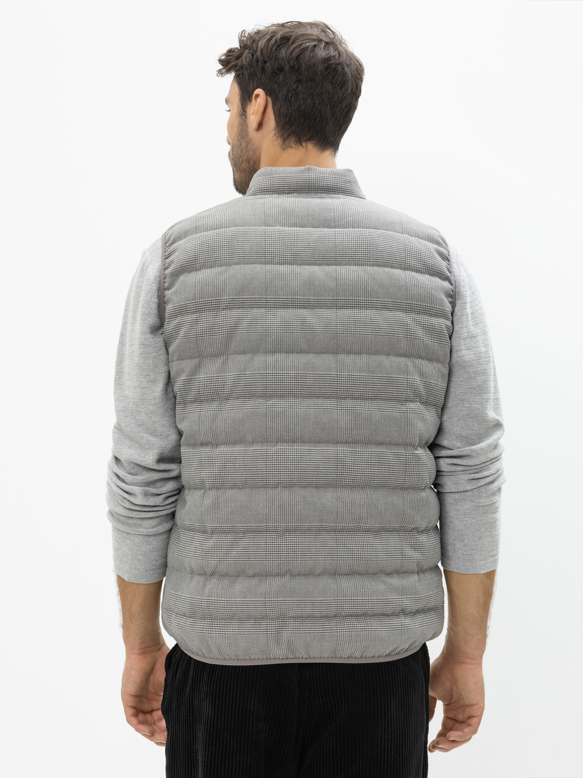 Ein Mann, der von sich wegschaut, trägt eine hellgraue Steppweste über einem grauen langärmeligen Hemd und eine schwarze Hose und steht vor einem schlichten weißen Hintergrund.
