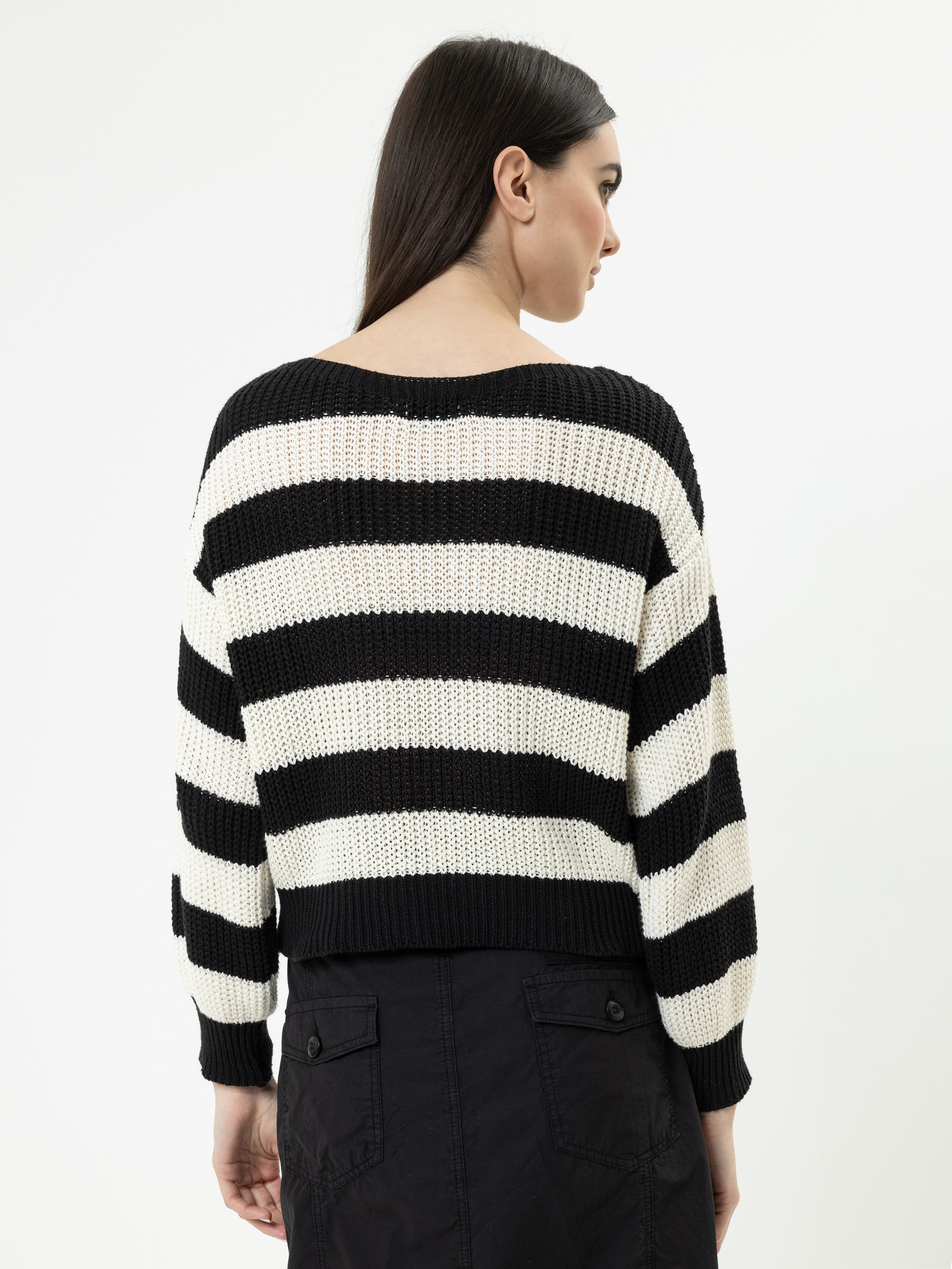 Eine Frau mit langen dunklen Haaren, von hinten gesehen, trägt einen schwarz-weiß horizontal gestreiften Strickpullover und einen schwarzen Rock vor einem schlichten weißen Hintergrund.