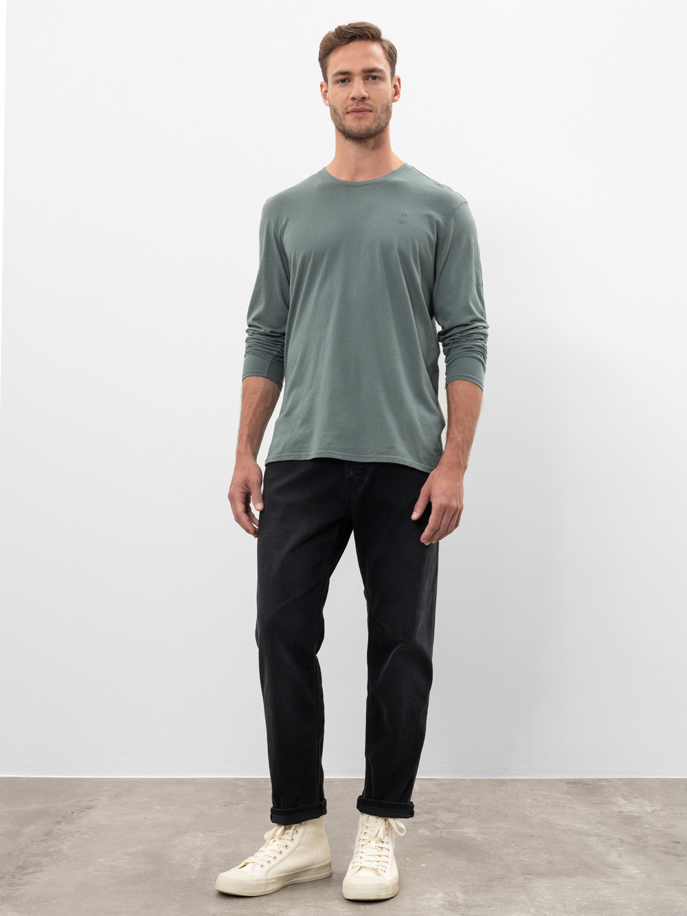 Ein Mann steht vor einer schlichten weißen Wand. Er trägt ein salbeigrünes langärmeliges Hemd, schwarze Jeans und cremefarbene Turnschuhe. Er blickt nach vorne und hat einen neutralen Gesichtsausdruck.