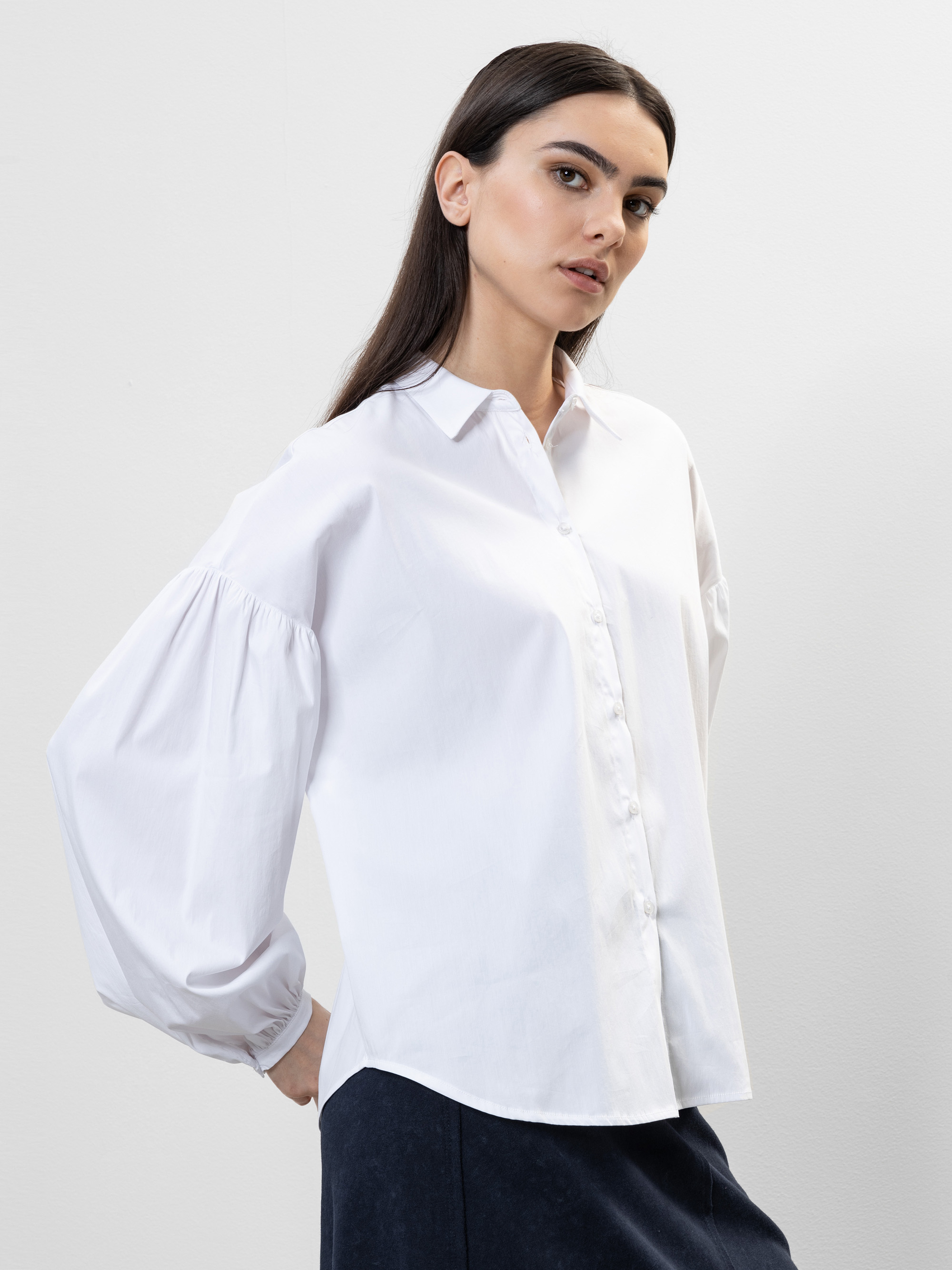 Eine Frau mit langen dunklen Haaren trägt eine locker sitzende weiße Button-up-Bluse mit Puffärmeln und eine dunkle Hose. Sie steht vor einem schlichten hellen Hintergrund und schaut leicht zur Seite.