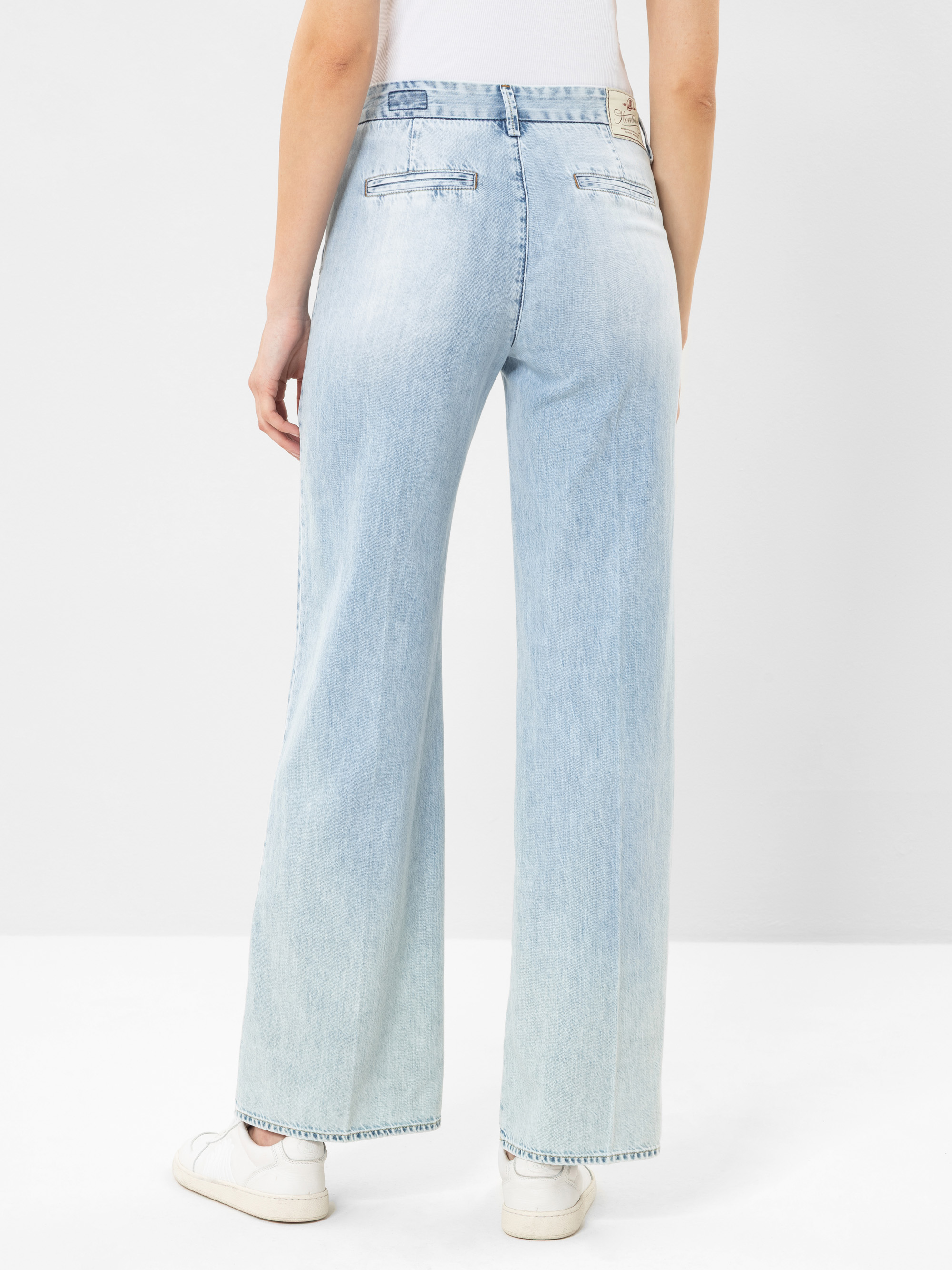 Eine Person, die hellblaue Jeans mit weitem Bein und weiße Turnschuhe trägt, wird von hinten gezeigt und steht vor einem schlichten weißen Hintergrund. Die Jeans hat Leistentaschen und eine hohe Taille.