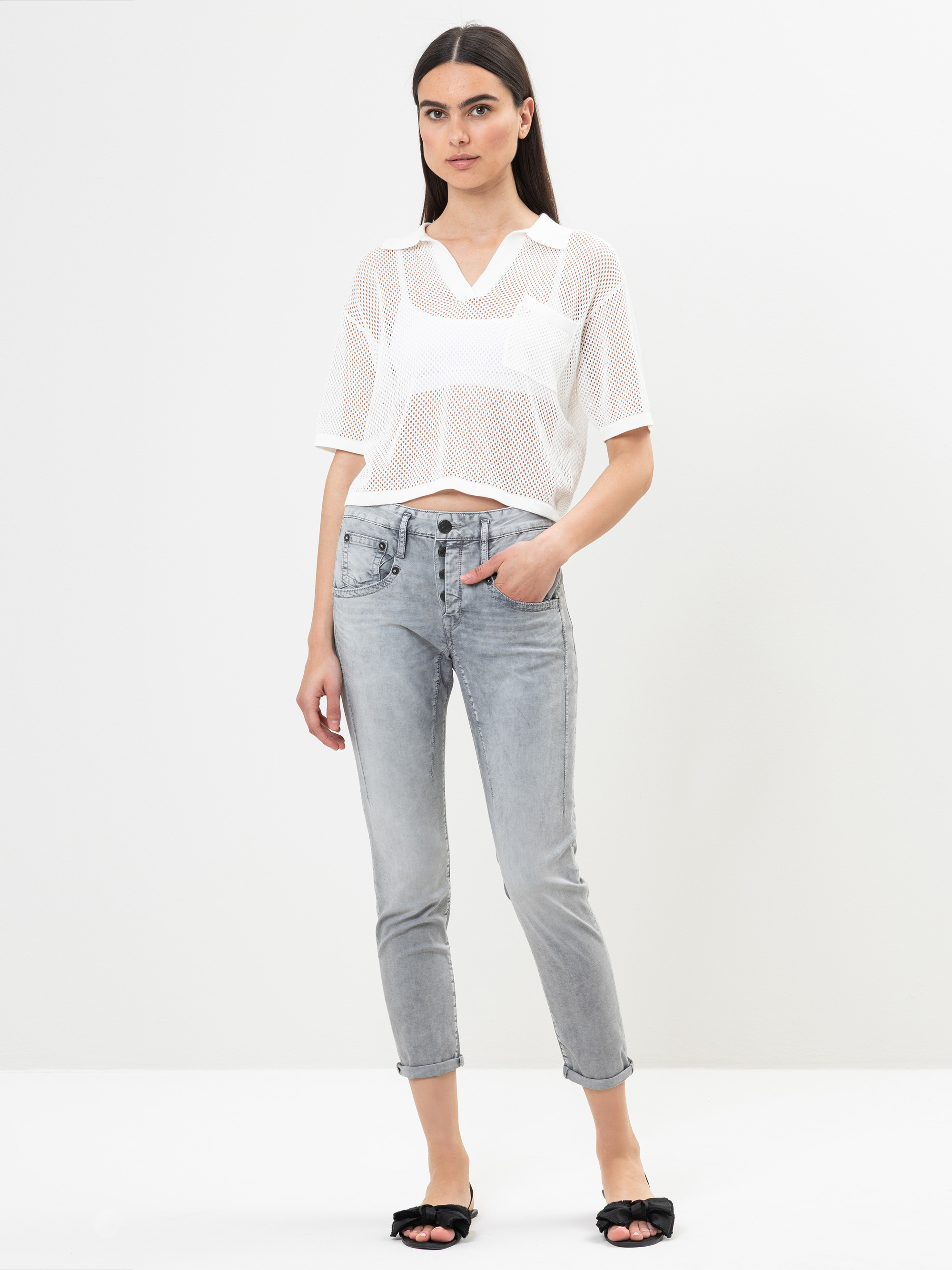 Eine Frau steht vor einem schlichten weißen Hintergrund und trägt ein durchsichtiges weißes Mesh-Top über einem weißen Bralette, hellgraue, kurze Jeans und schwarze Slide-Sandalen. Sie hat glattes dunkles Haar und einen neutralen Gesichtsausdruck, ei