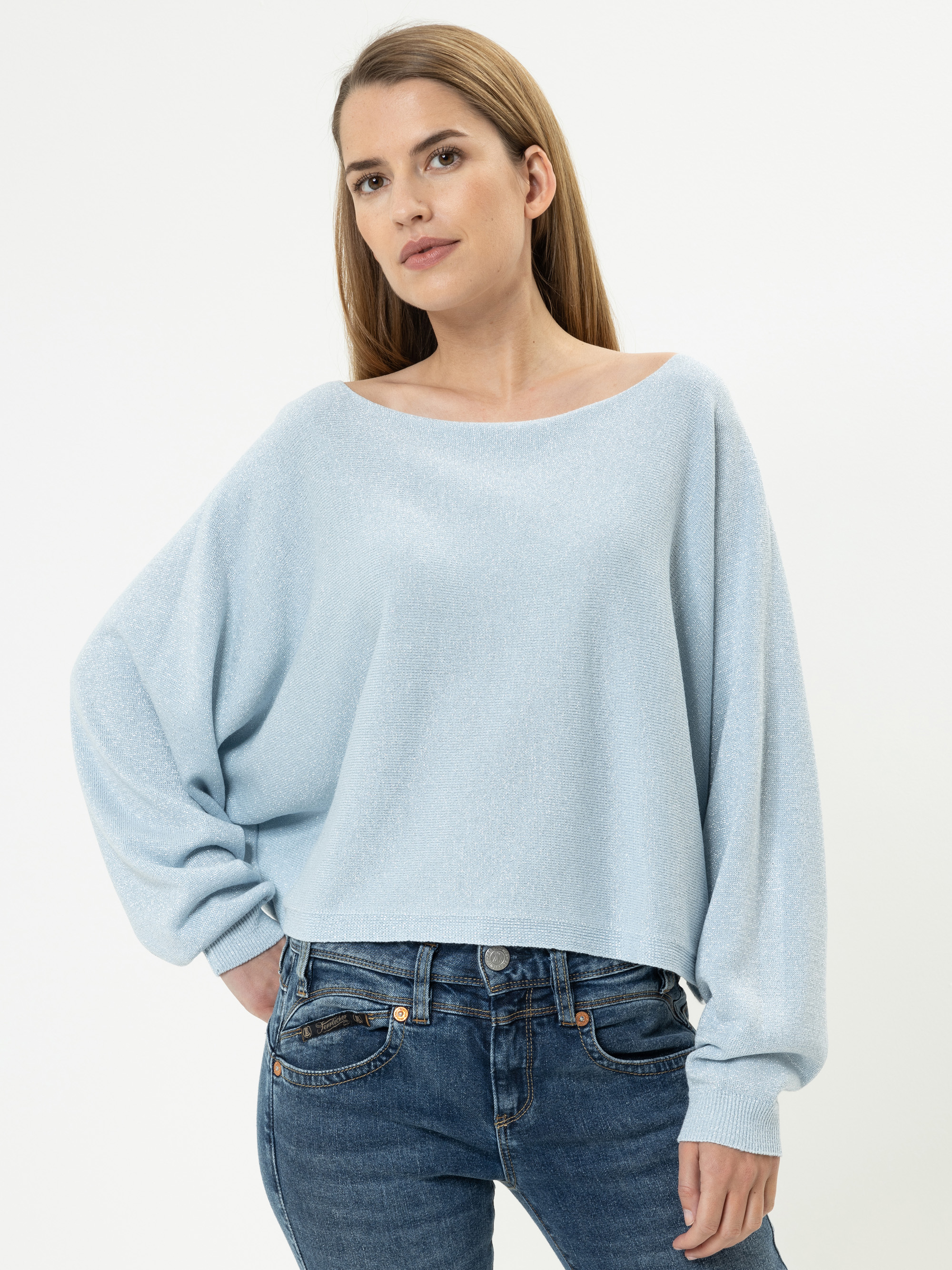 Eine Frau mit langem, glattem braunem Haar trägt einen lockeren, hellblauen Pullover mit weiten Ärmeln und blaue Jeans. Sie steht vor einem schlichten weißen Hintergrund und hat eine Hand in die Hüfte gestemmt.