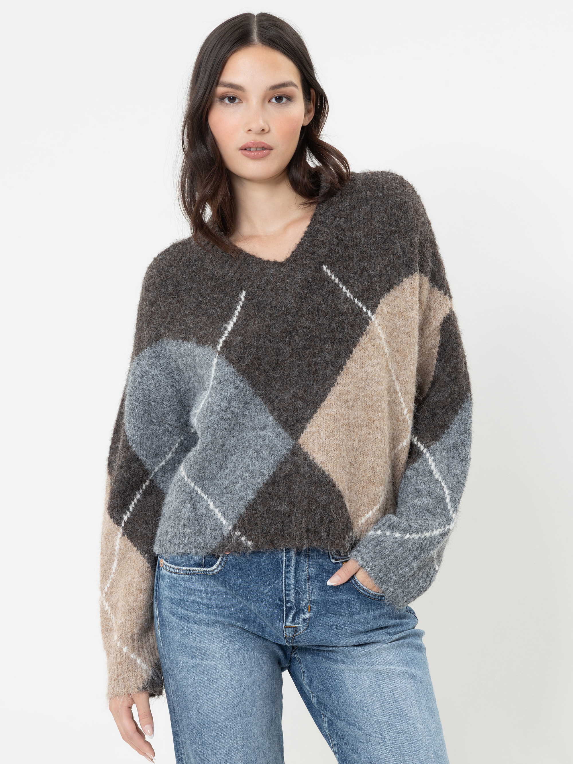 Eine Frau mit langen dunklen Haaren, die einen flauschigen Pullover in den Farben Braun, Grau und Beige mit weißen Streifen sowie eine blaue Jeans trägt und vor einem schlichten weißen Hintergrund steht, wobei sie eine Hand in die Tasche gesteckt hat.