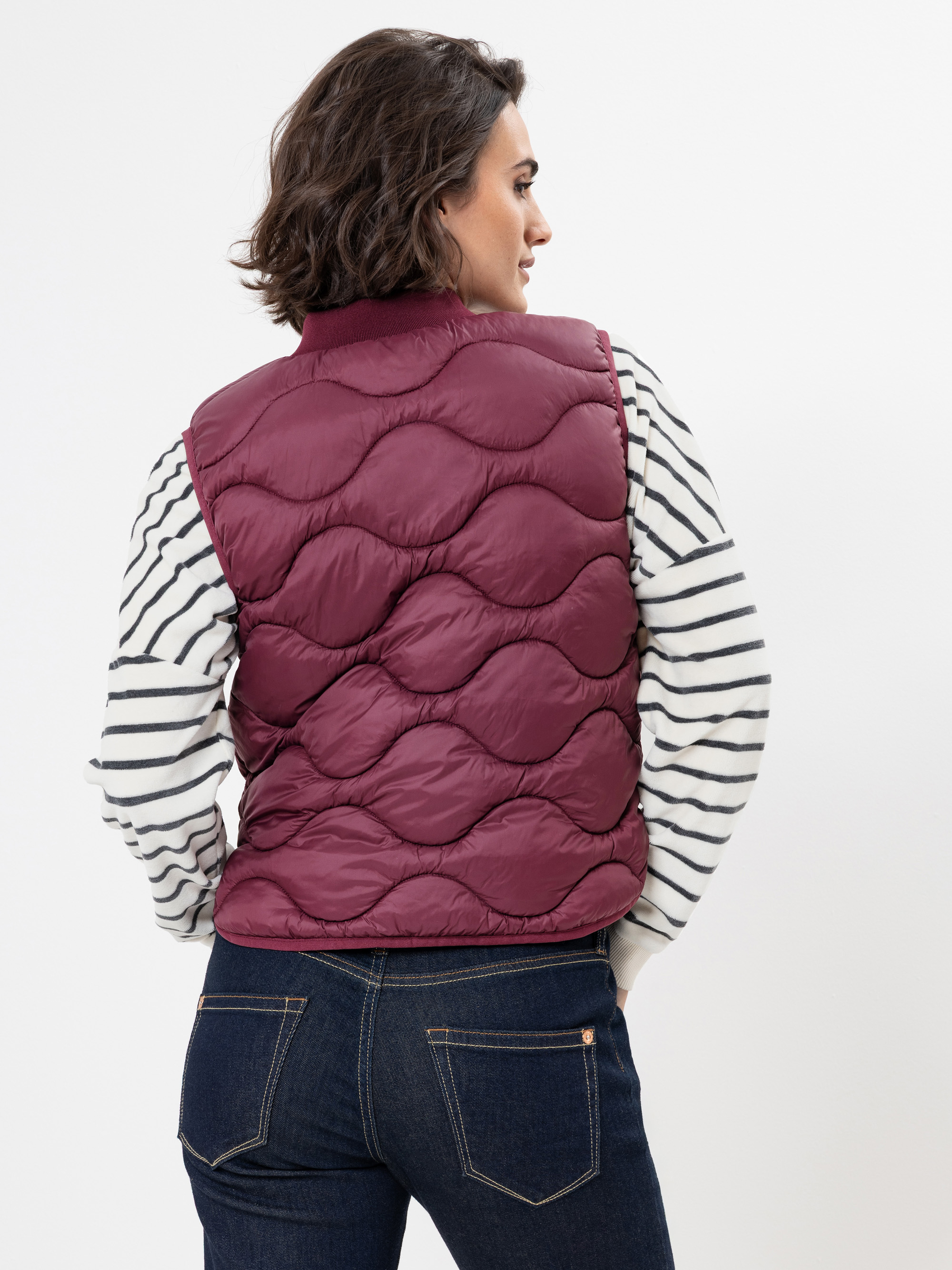 Eine Frau steht mit dem Gesicht zur Wand, trägt eine kastanienbraune Steppweste über einem weiß-schwarz gestreiften langärmeligen Hemd und dunkelblaue Jeans vor einem schlichten weißen Hintergrund.