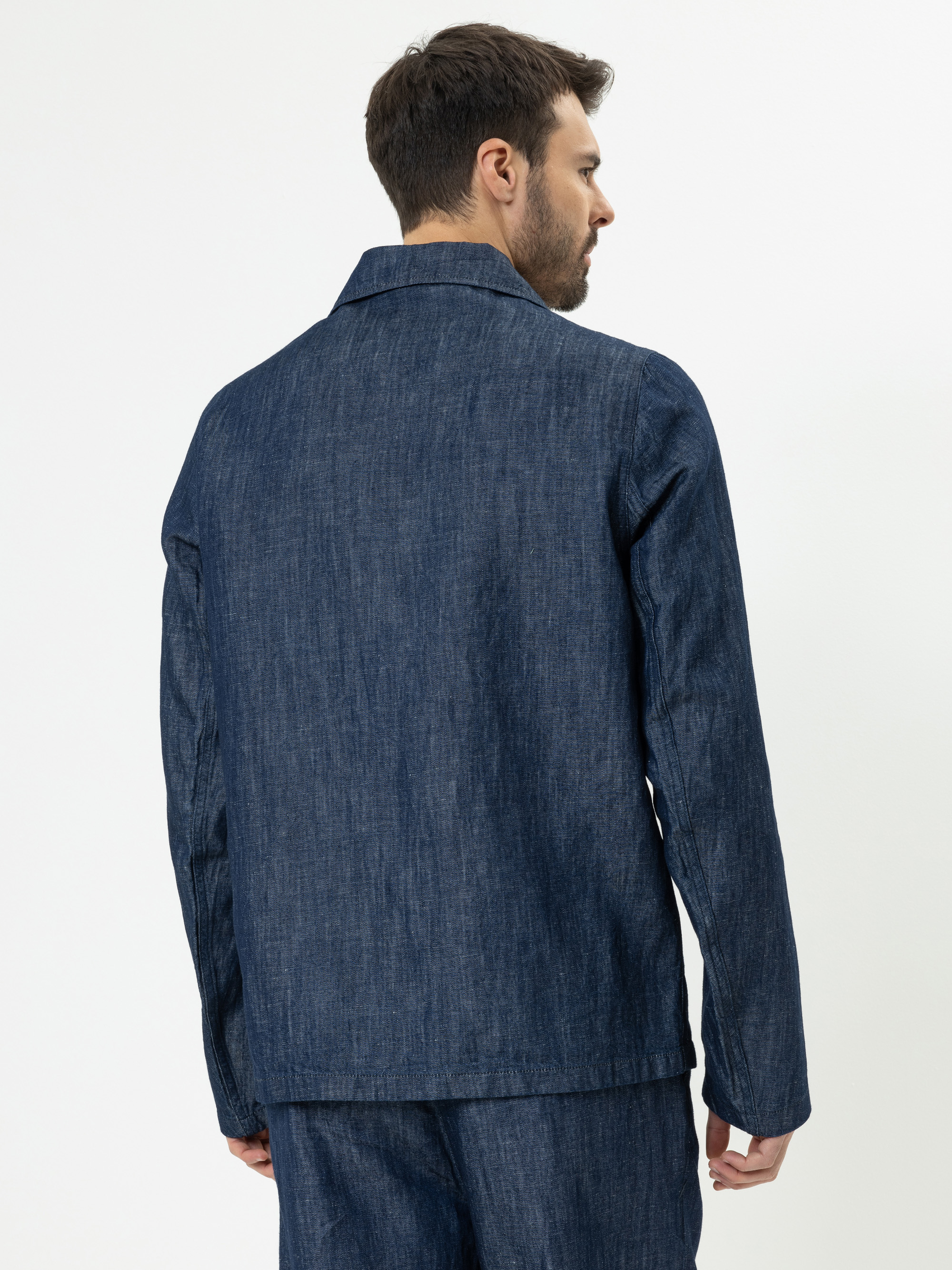 Ein Mann mit kurzen braunen Haaren wird von hinten gezeigt, der eine dunkelblaue Jeansjacke und eine dazu passende Hose trägt und vor einem schlichten weißen Hintergrund steht.