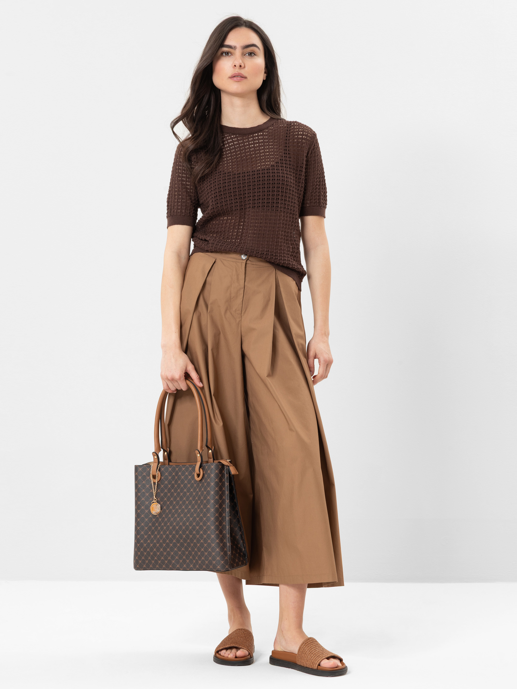 Eine Frau steht vor einem unifarbenen Hintergrund und trägt ein braunes, kurzärmeliges Strickoberteil, eine weite, hellbraune Culotte-Hose, braune Gleitsandalen und eine braun gemusterte Tragetasche.