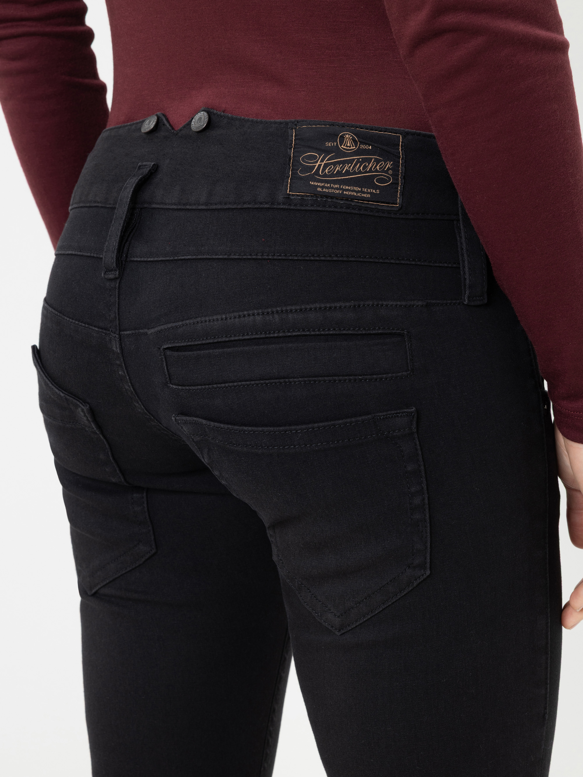 Person, die ein kastanienbraunes Oberteil und eine schwarze Herrlicher-Jeans trägt; die Aufnahme zeigt die Person von hinten, um die zahlreichen Gesäßtaschen und das braune Markenetikett am Bund zu zeigen.
