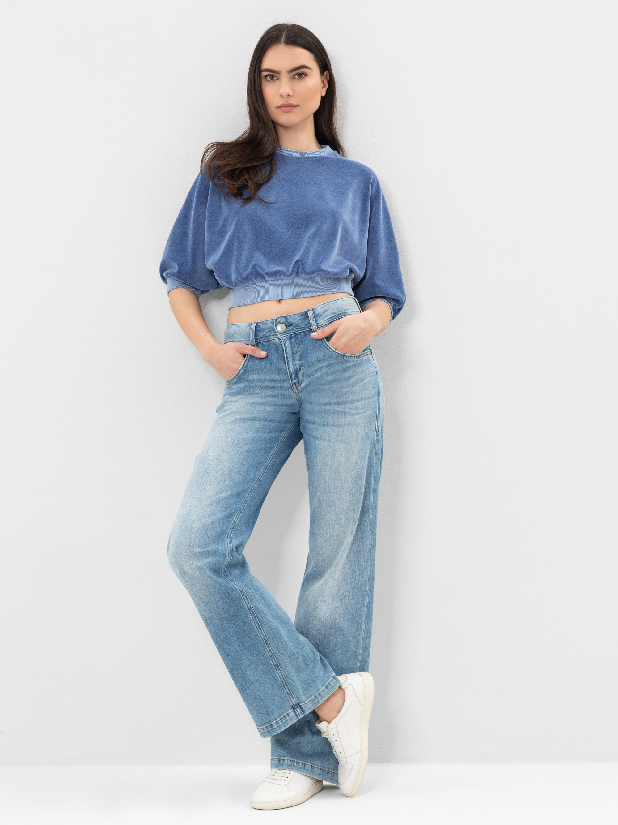 Frau, die vor einem schlichten, hellen Hintergrund steht. Sie trägt ein kurzärmeliges Crop-Top aus blauem Samt, hellblaue Jeans mit hoher Taille und weiten Beinen sowie weiße Turnschuhe. Sie hat langes dunkles Haar und die Hände in den Taschen.