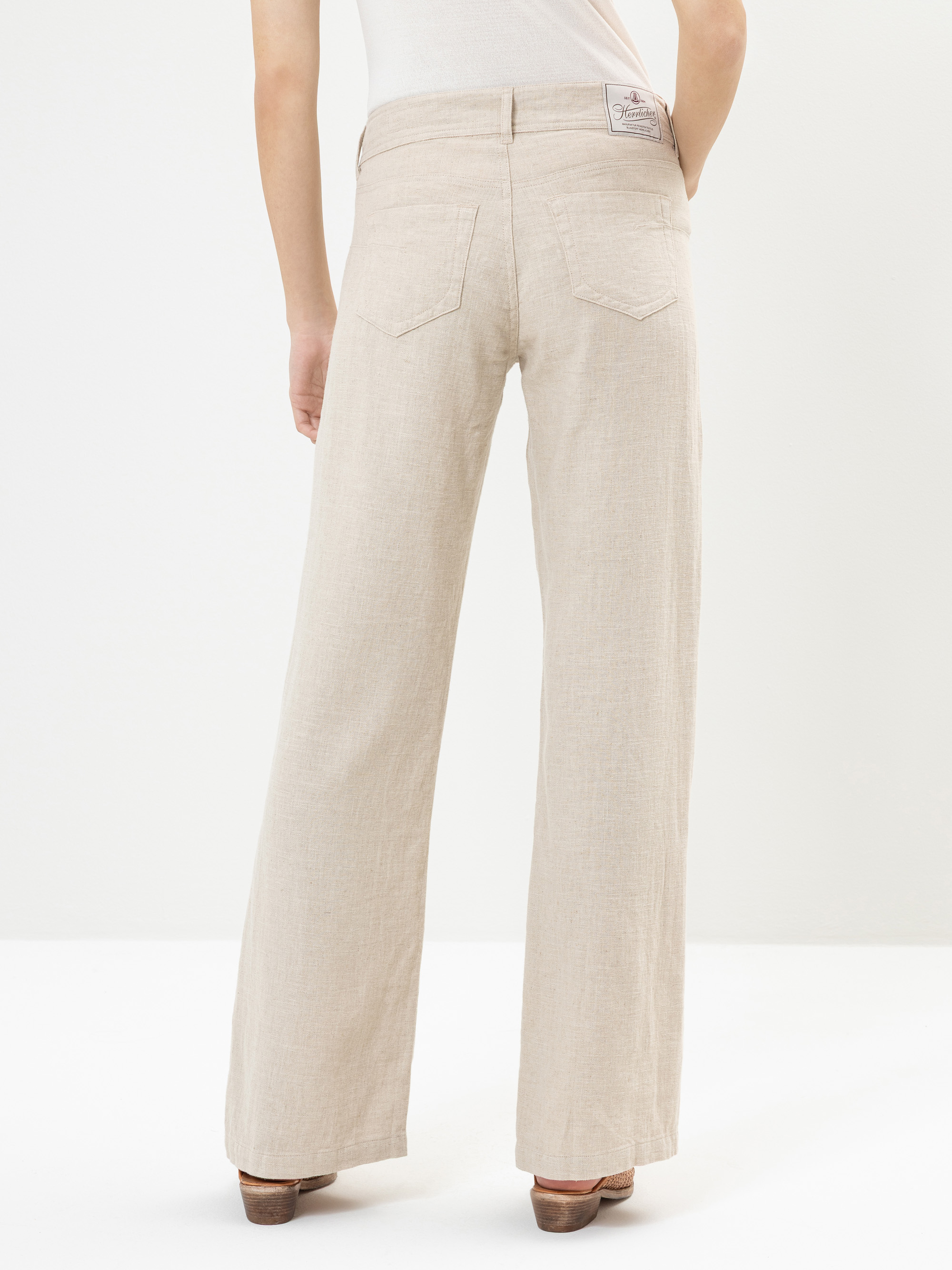 Eine Person, die eine cremefarbene Hose mit weitem Bein und braune Schuhe mit Absätzen trägt, wird von hinten gezeigt und steht vor einem schlichten hellen Hintergrund.