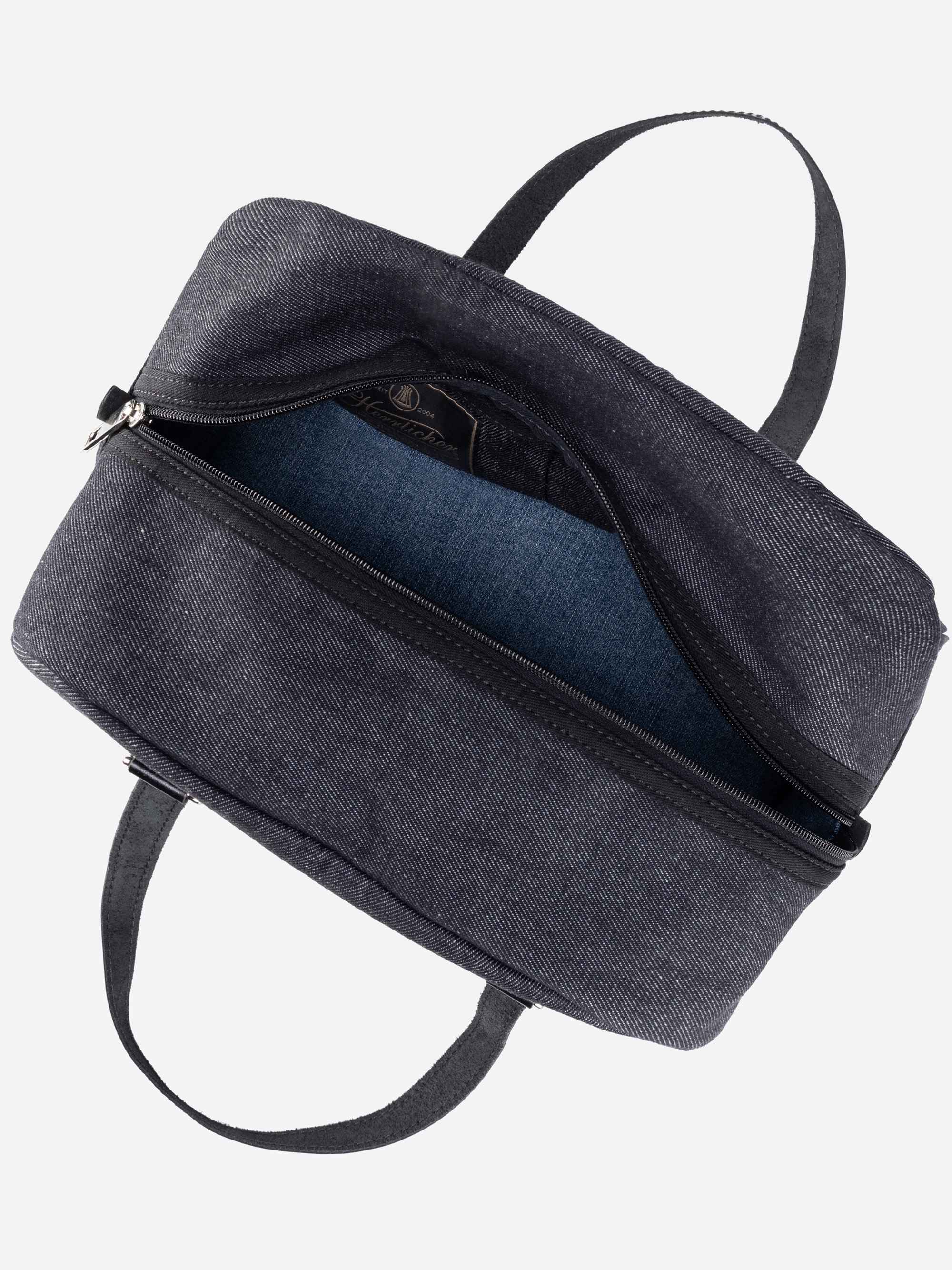 Eine Tragetasche aus dunklem Denim mit schwarzen Griffen und einer Öffnung mit Reißverschluss, von oben gezeigt. Die Tasche ist teilweise geöffnet und offenbart ein blaues Stofffutter und eine Innentasche.