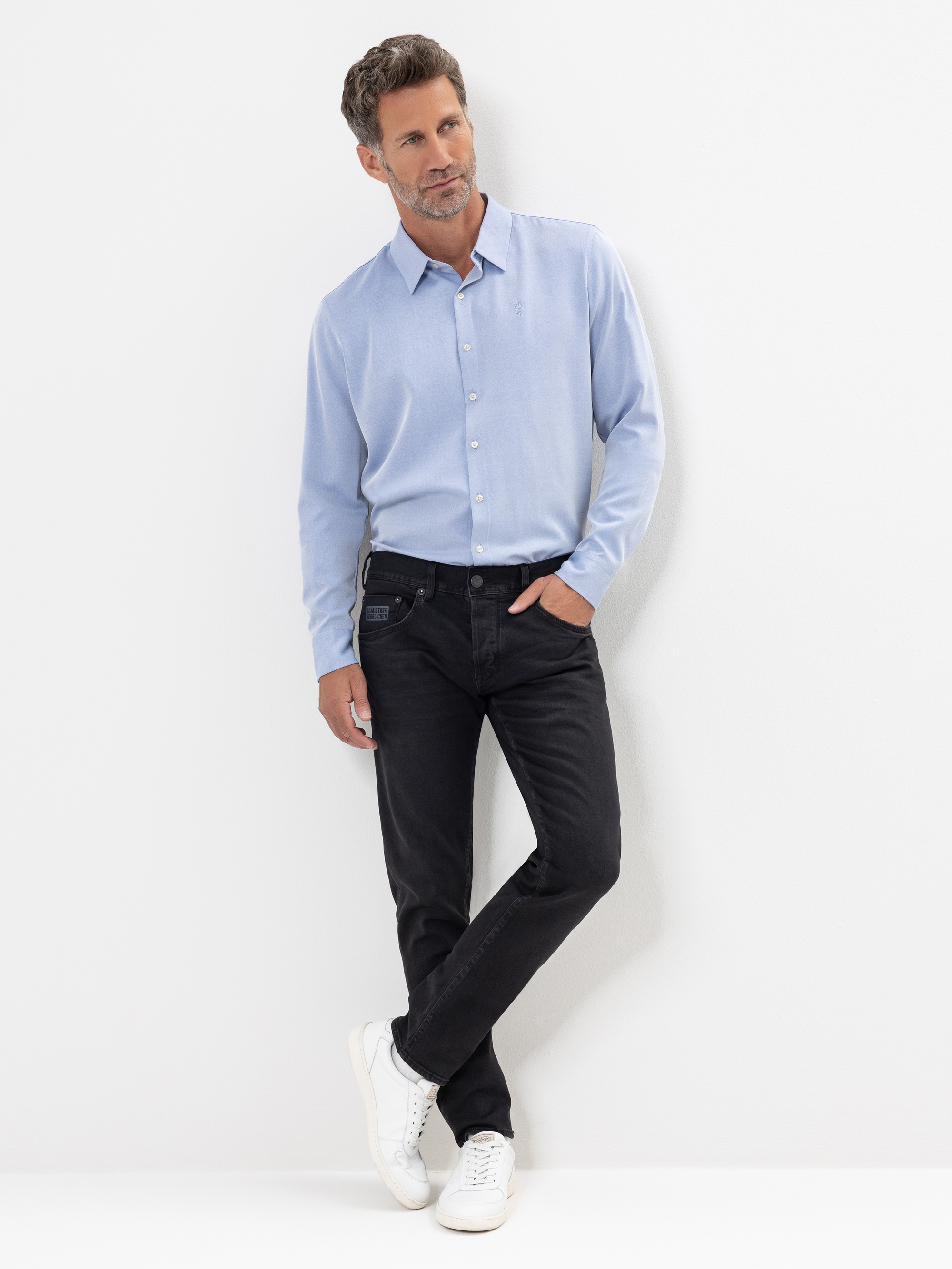 Ein Mann steht an einer weißen Wand und trägt ein hellblaues Button-up-Hemd, schwarze Jeans und weiße Turnschuhe. Er hat eine Hand in der Hosentasche, die Beine am Knöchel gekreuzt und schaut leicht zur Seite.