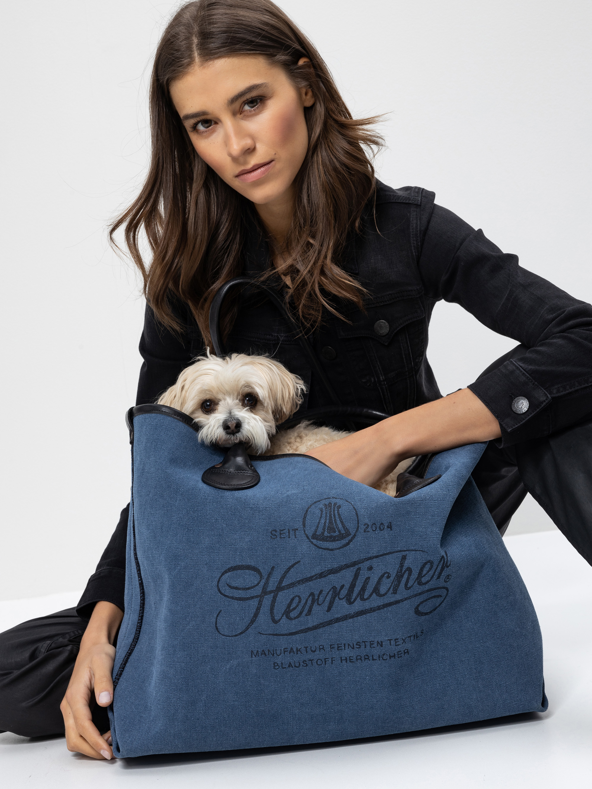 Eine Frau in schwarzer Kleidung sitzt auf dem Boden und hält eine große blaue Tasche mit der Aufschrift Herrlicher in der Hand. Ein kleiner beiger Hund sitzt bequem in der Tasche und lugt heraus.