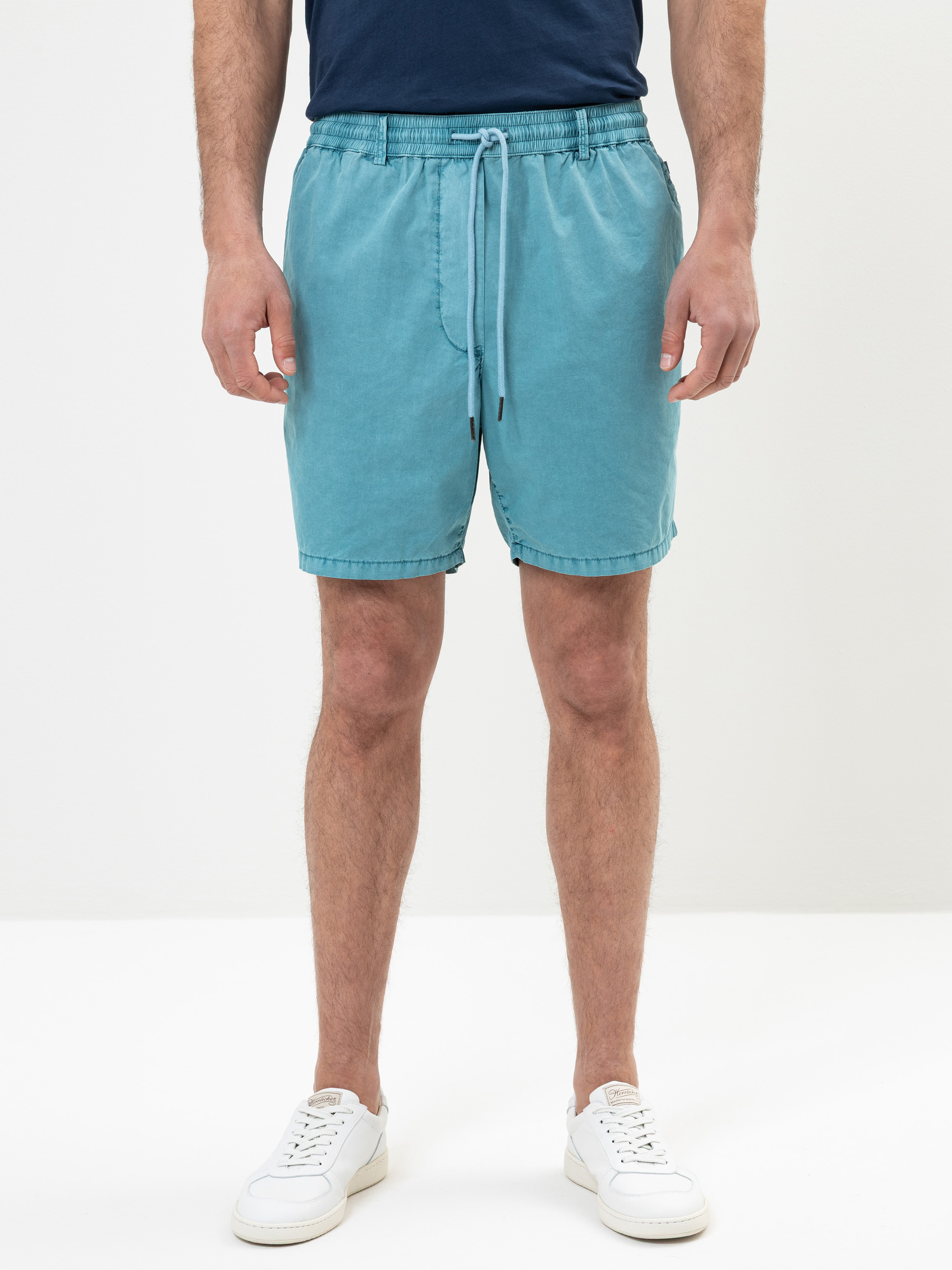 Eine Person in aquablauen Shorts, einem marineblauen T-Shirt und weißen Turnschuhen, die vor einem schlichten weißen Hintergrund steht. Das Bild ist von den Schultern abwärts beschnitten.
