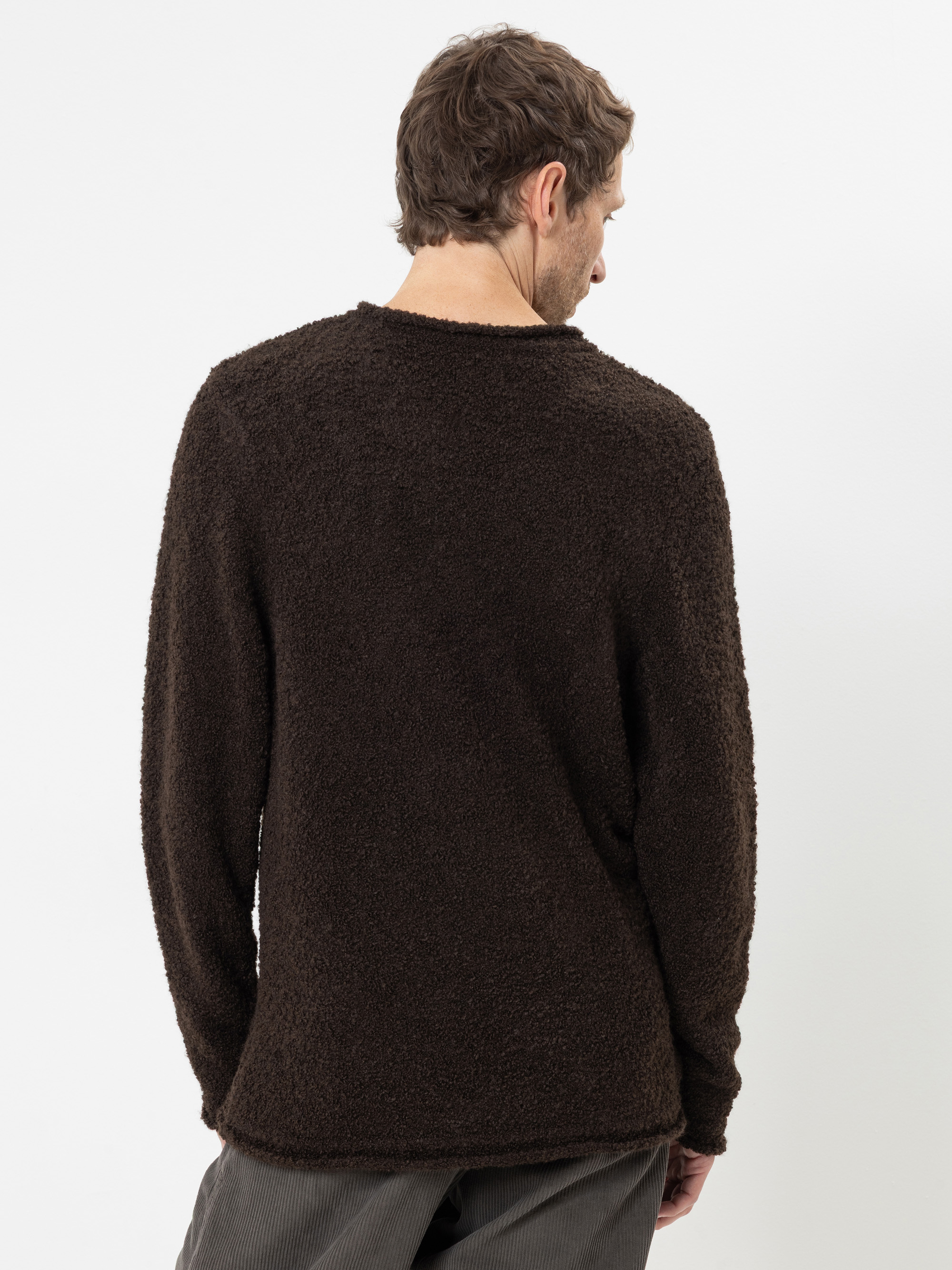 Vor einem schlichten weißen Hintergrund steht ein Mann mit dem Rücken zur Kamera; er trägt einen langärmeligen, dunkelbraunen, strukturierten Pullover und eine graue Hose.
